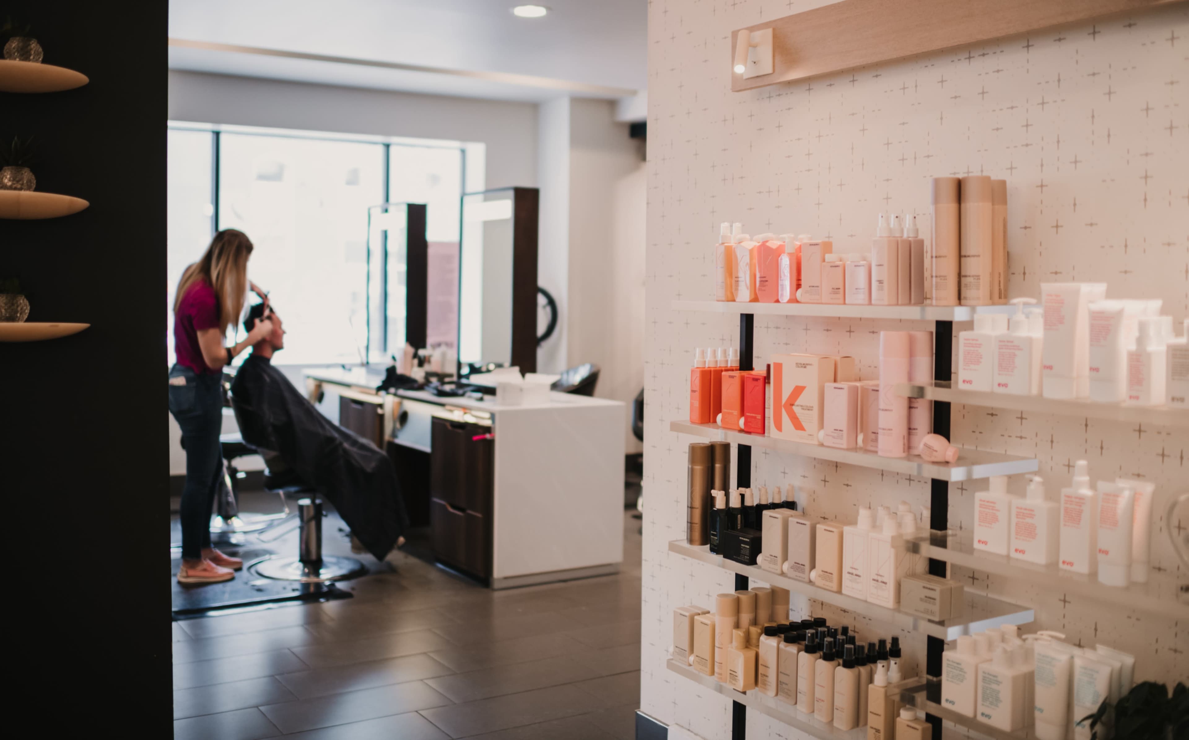 A person is seated in a salon chair while a stylist works on their hair, with a display of hair products on the wall nearby.