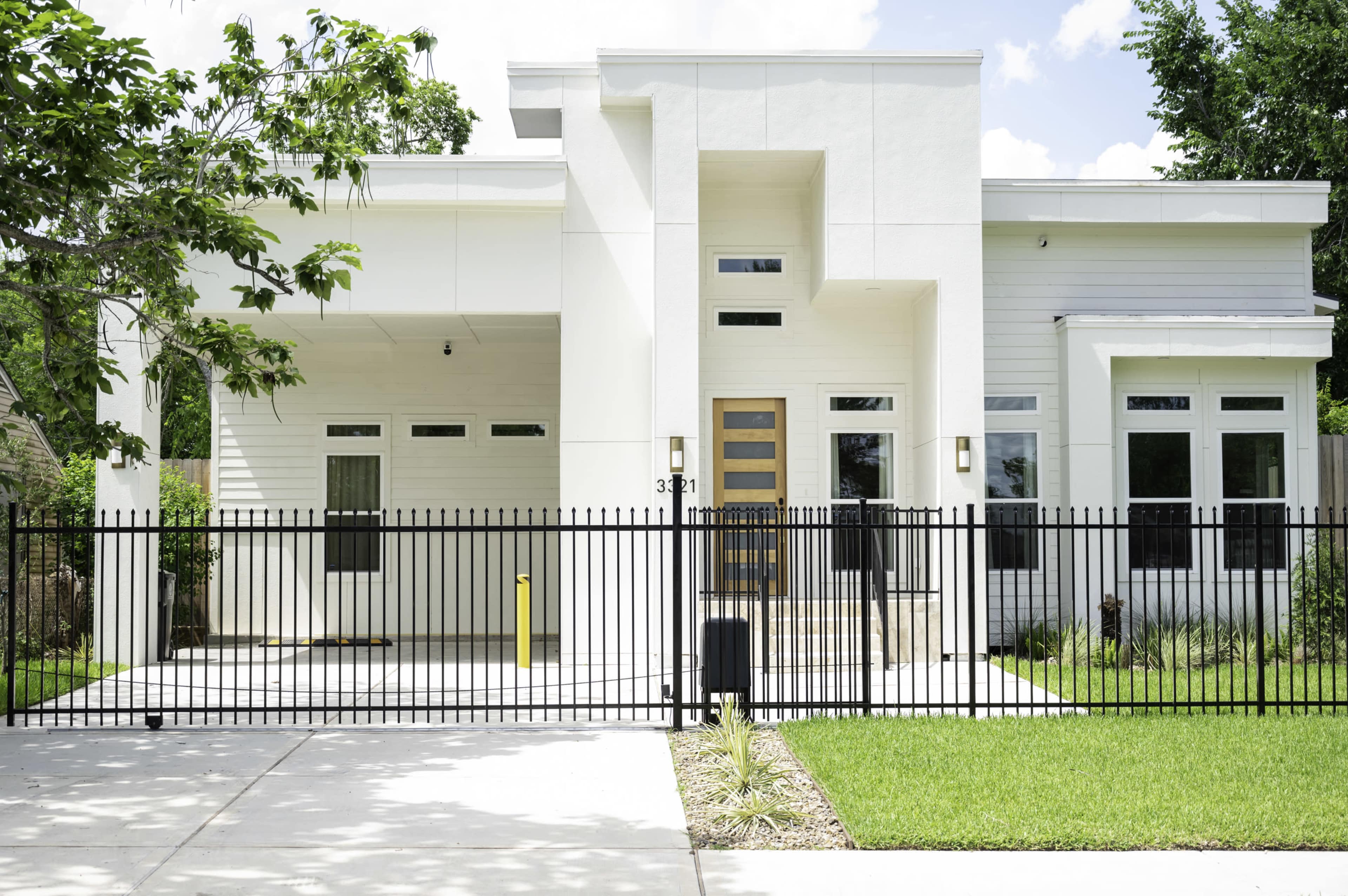 The image shows a modern single-story house with a white facade, large windows, and a black metal fence surrounding a landscaped front yard.