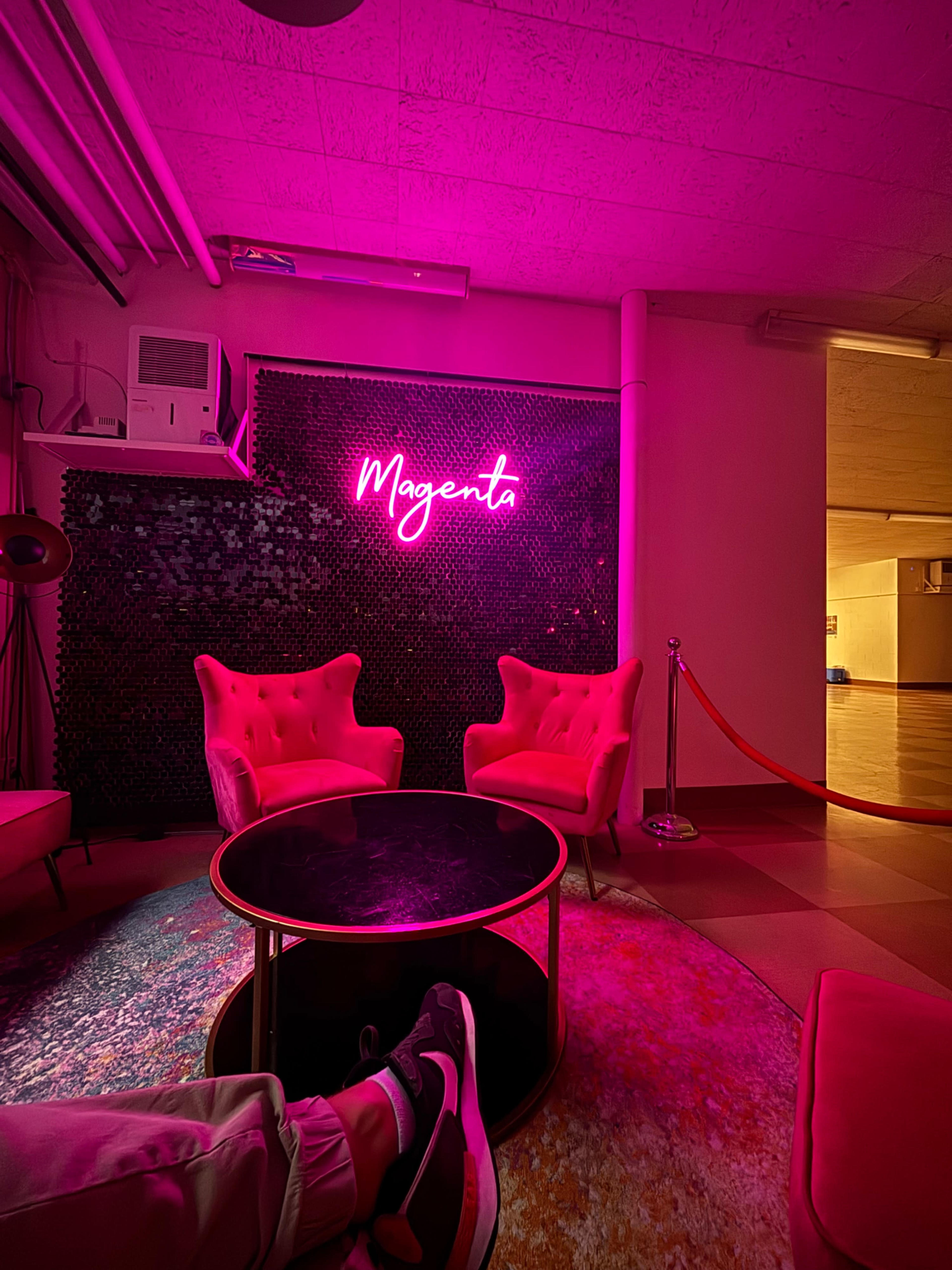 The image shows a dimly lit lounge area with pink neon signage reading "Magenta," featuring two plush pink chairs and a glass coffee table.