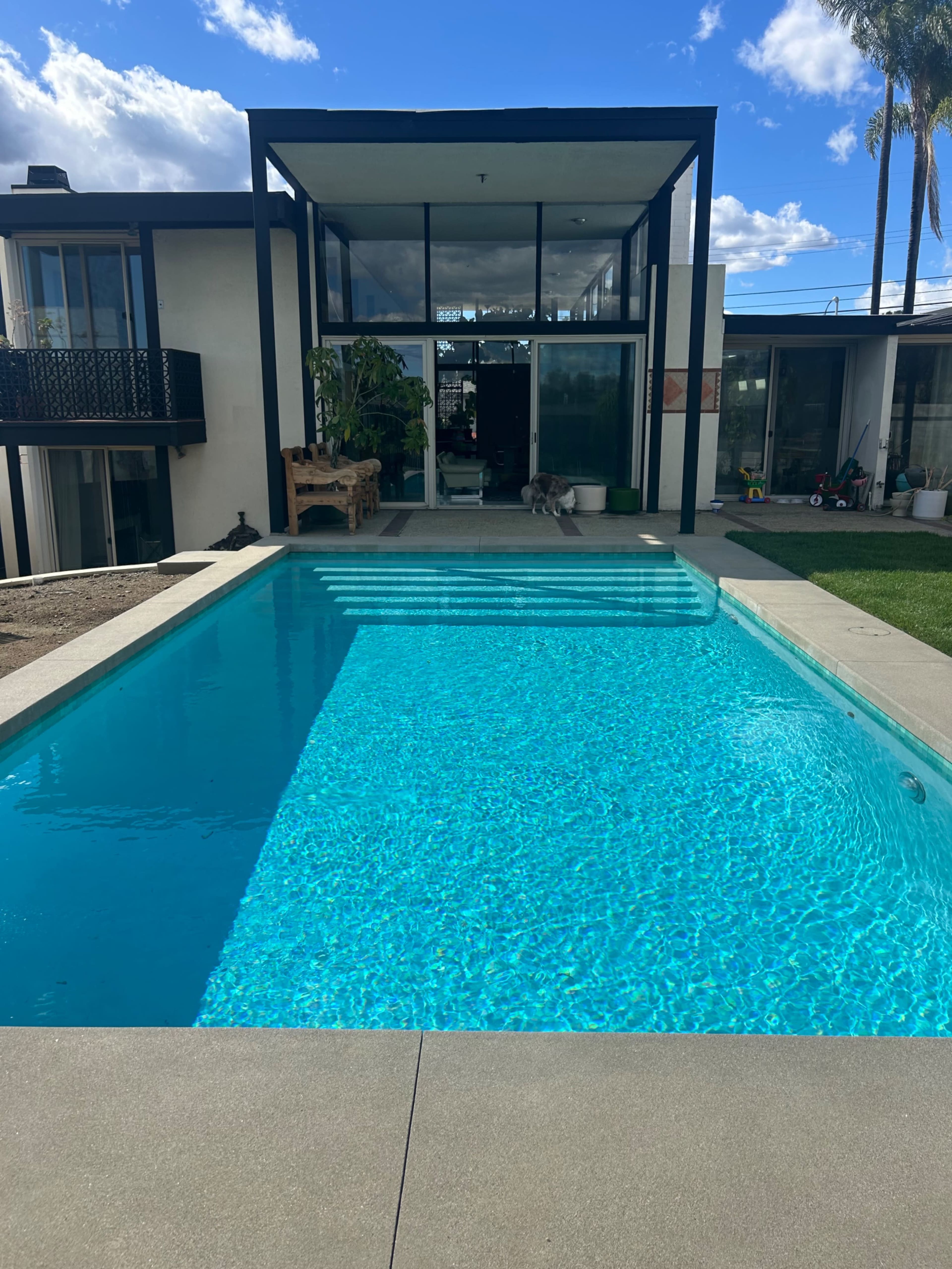 The image shows a modern home with a clear blue swimming pool in the foreground, leading up to a stylish structure with large windows and greenery.