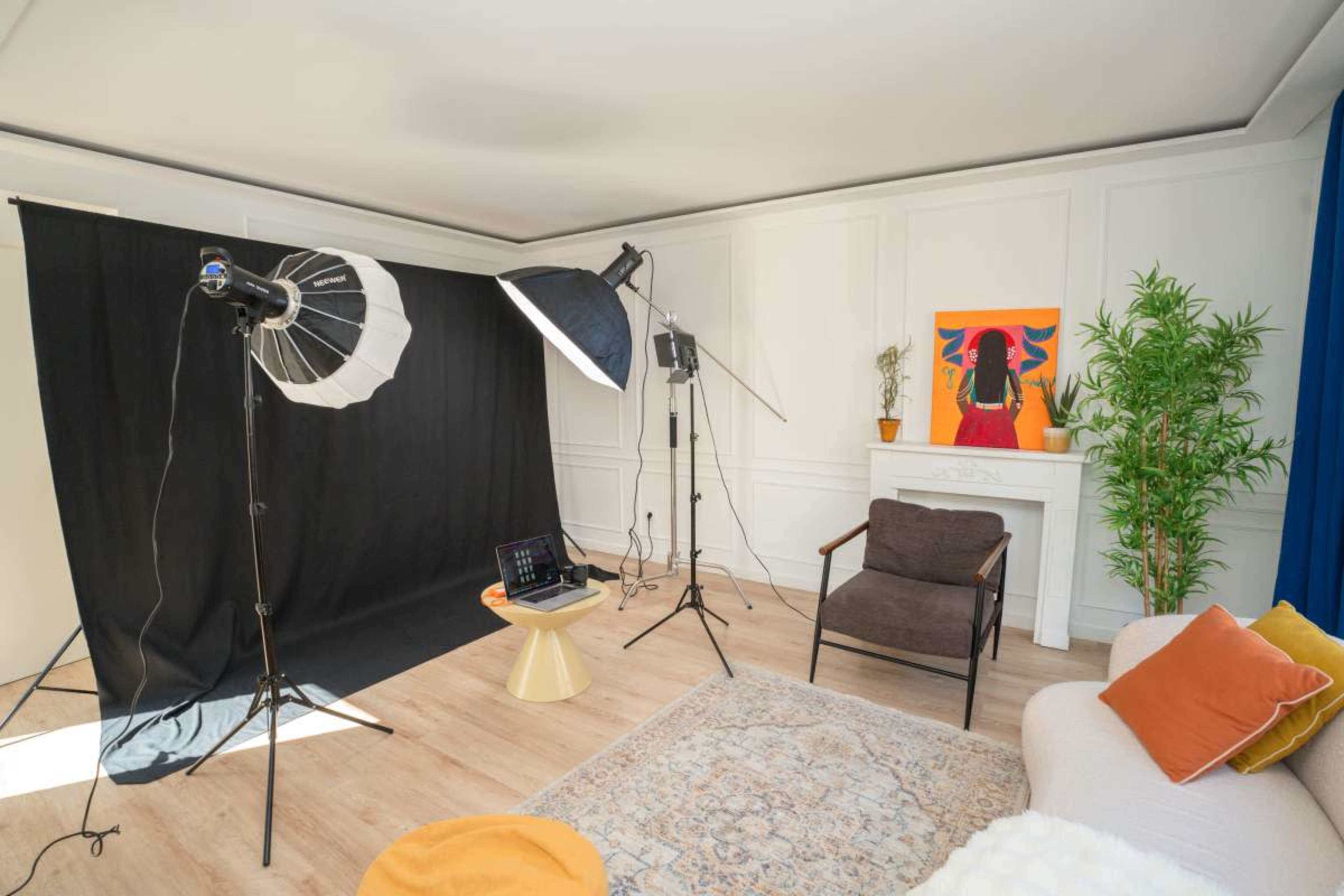 The image depicts a photography studio setup with a black backdrop, studio lights, a laptop on a side table, and a decorative plant next to a chair and a sofa.