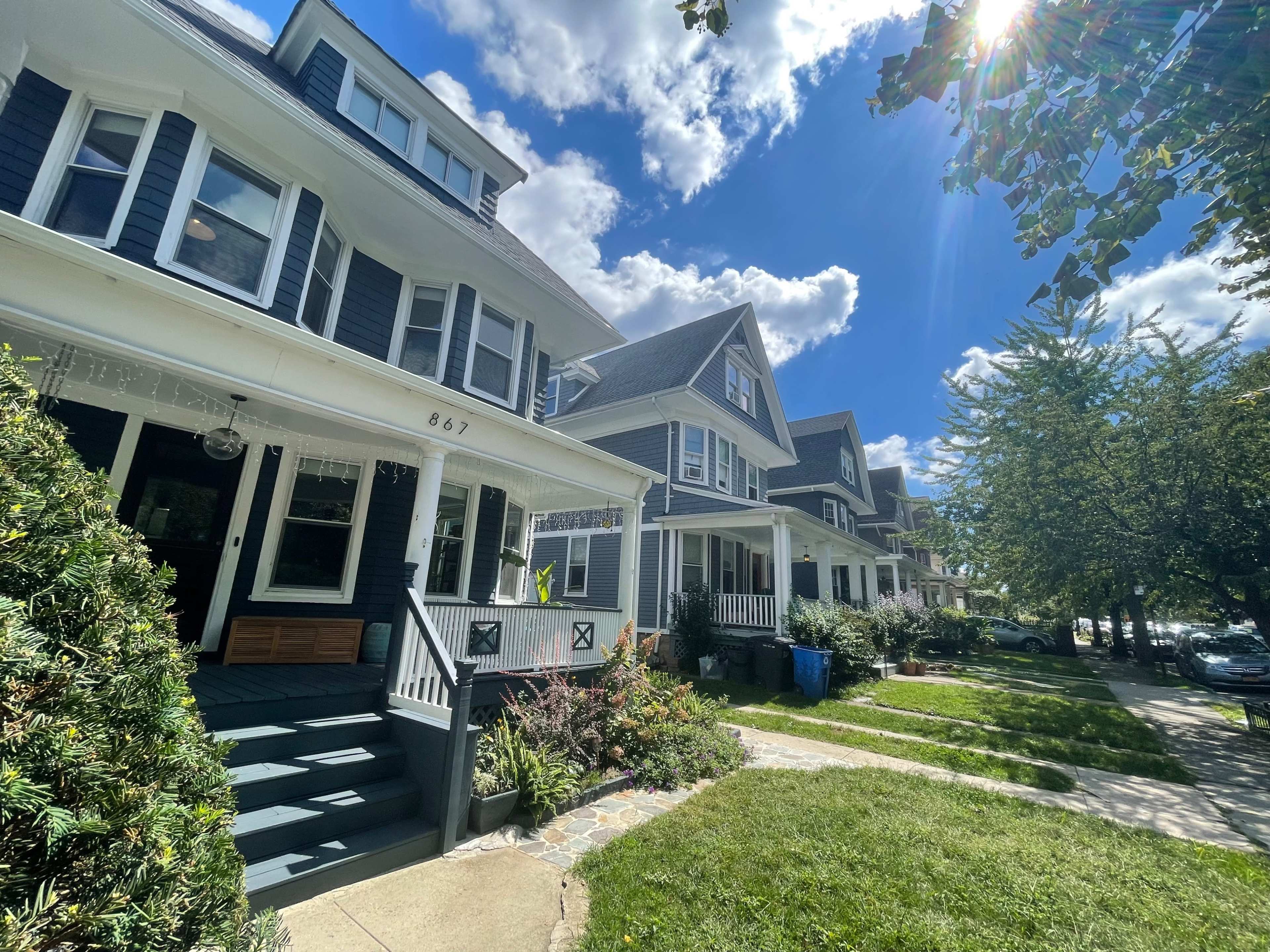 A row of traditional-style houses with porches line a sunny street, accompanied by neatly trimmed lawns and blue trash bins.