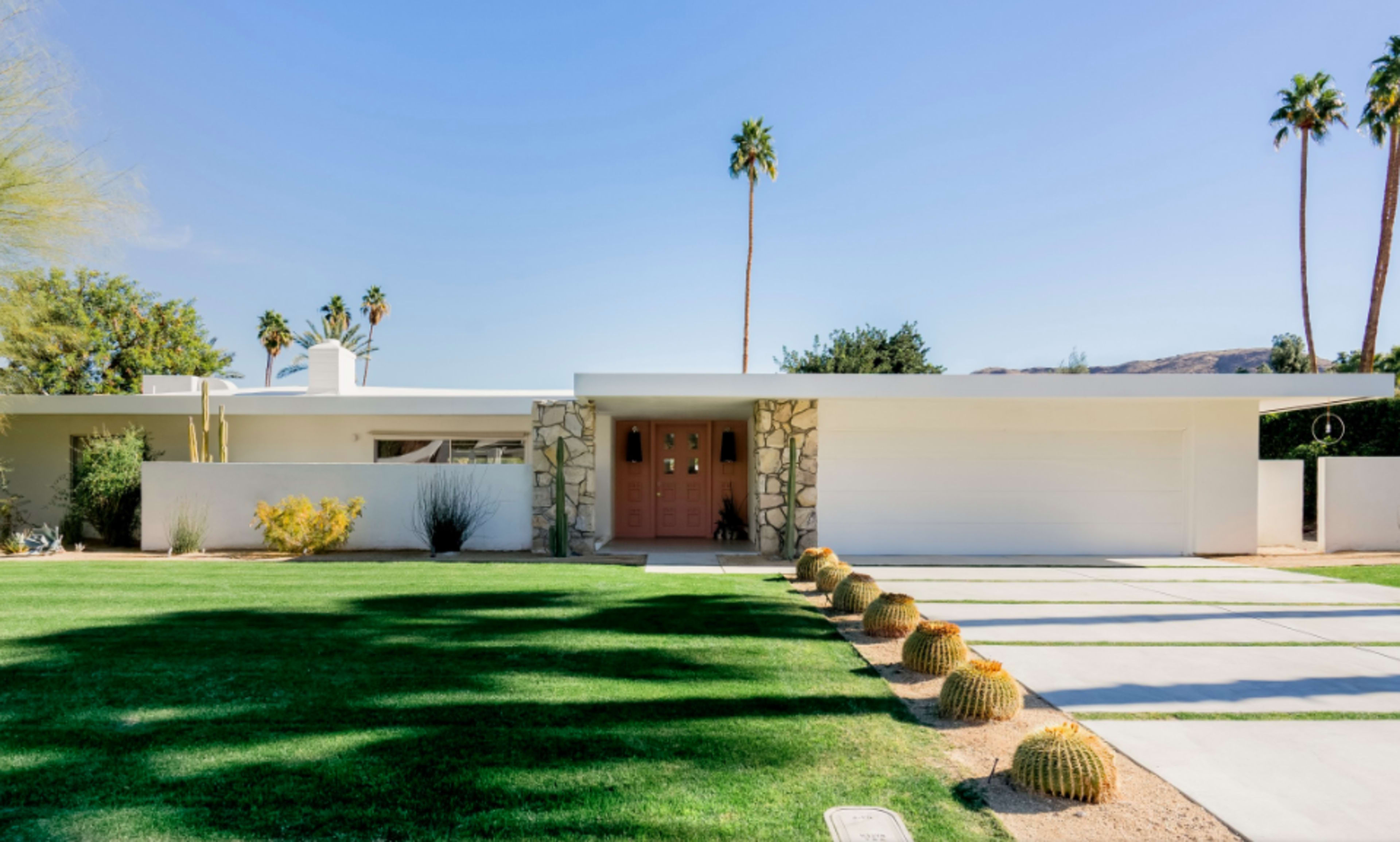 A modern single-story house with a stone-accented entrance, a large garage door, and landscaped front yard featuring cacti and palm trees.