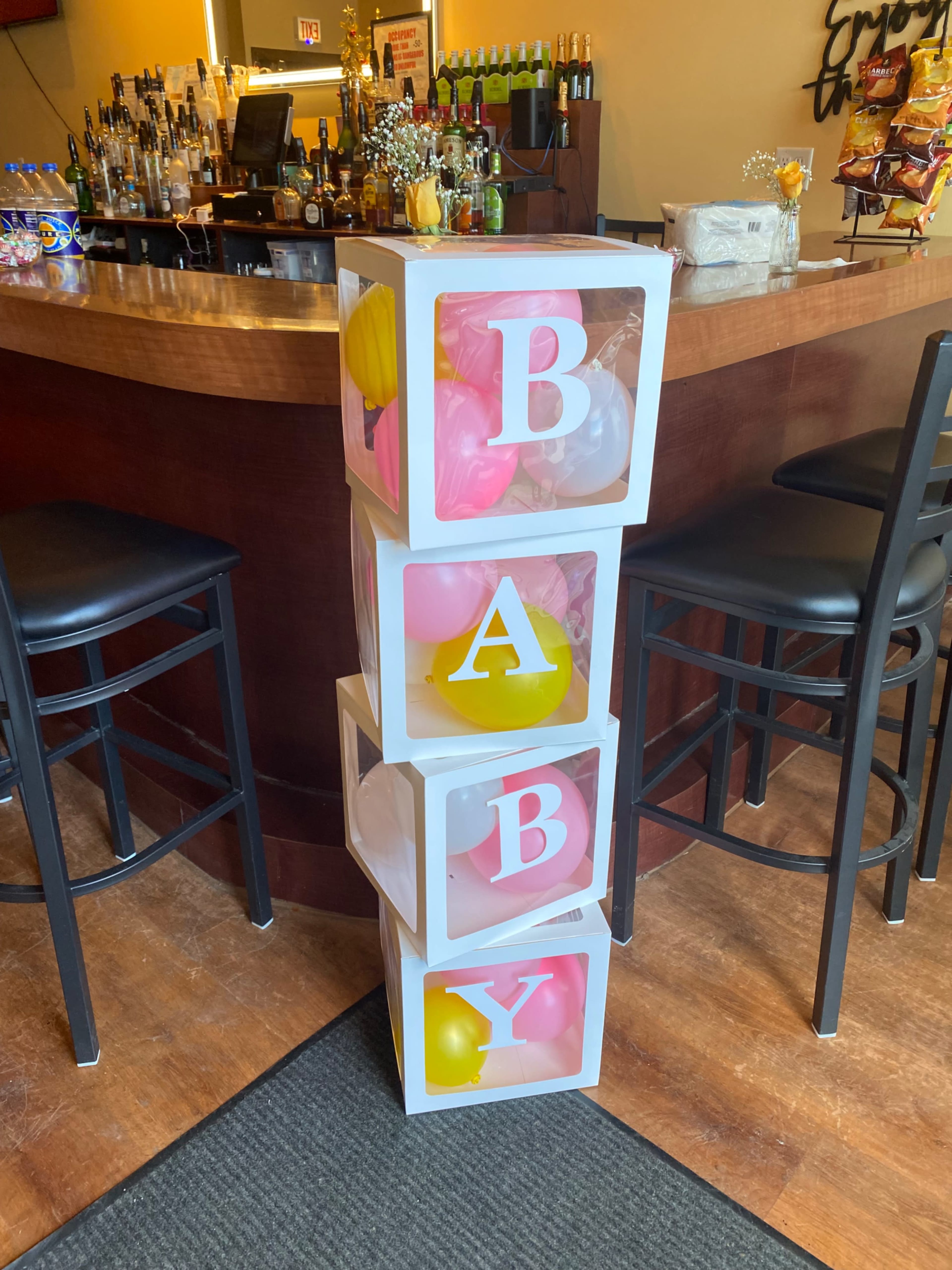 A tower of four boxes spelling "BABY," filled with colorful balloons, is positioned near a bar counter.
