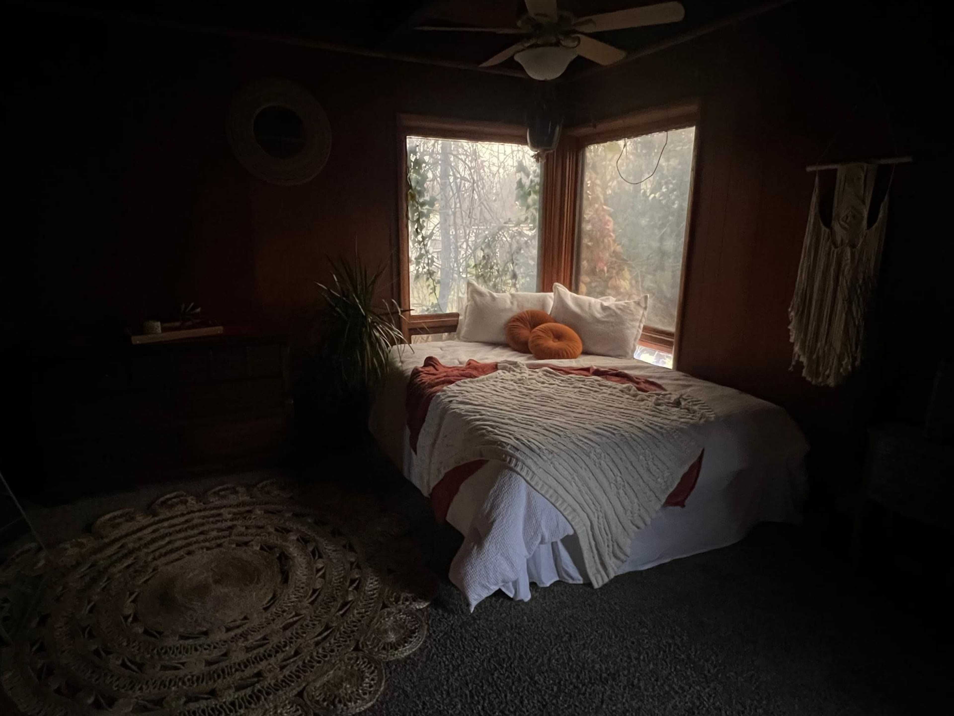 A cozy bedroom features a large bed with white bedding and orange accent pillows, surrounded by windows that let in soft, diffused light.