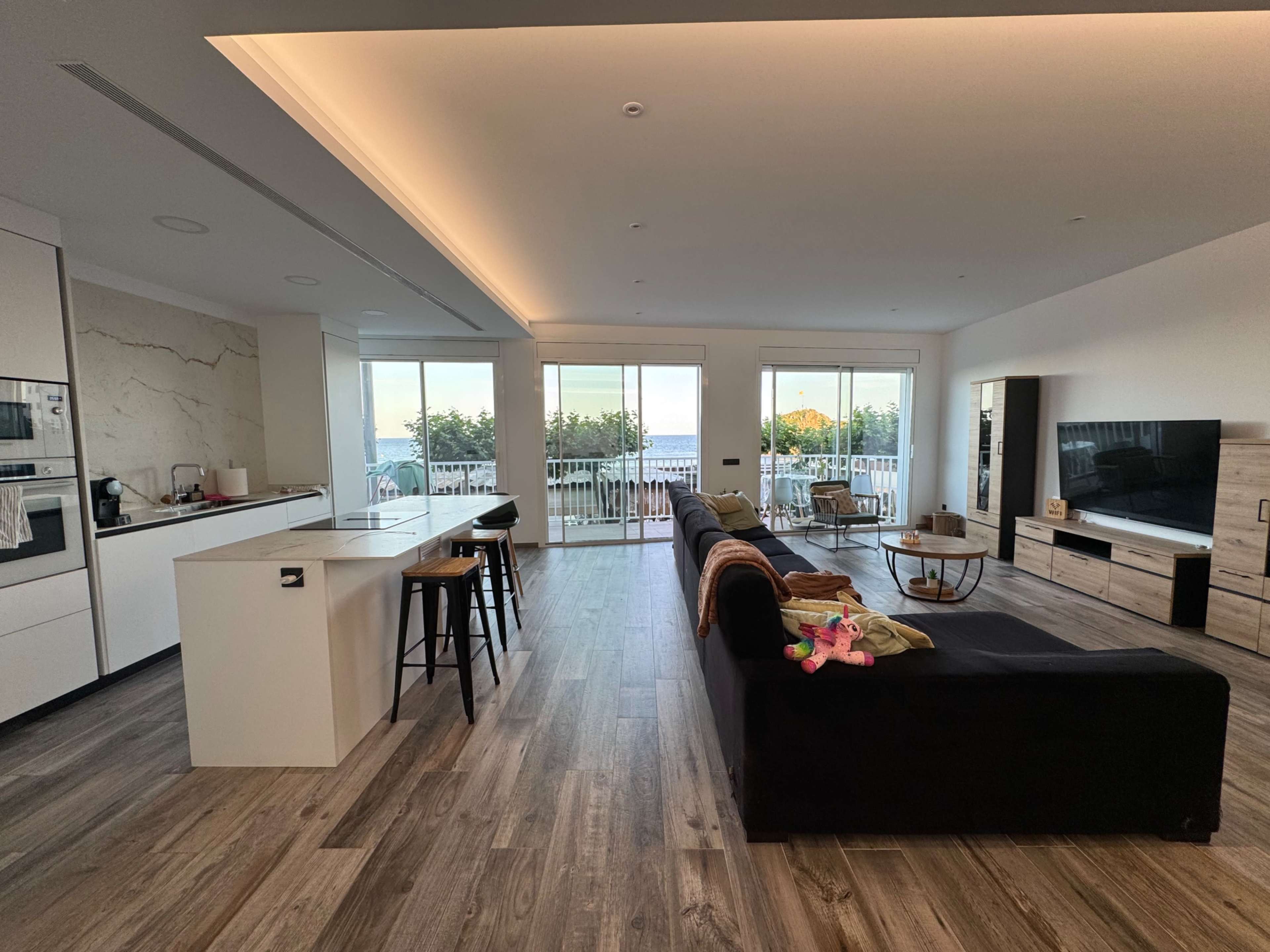 The image shows a modern living room connected to a kitchen area, featuring large windows that open to a balcony with views of greenery.