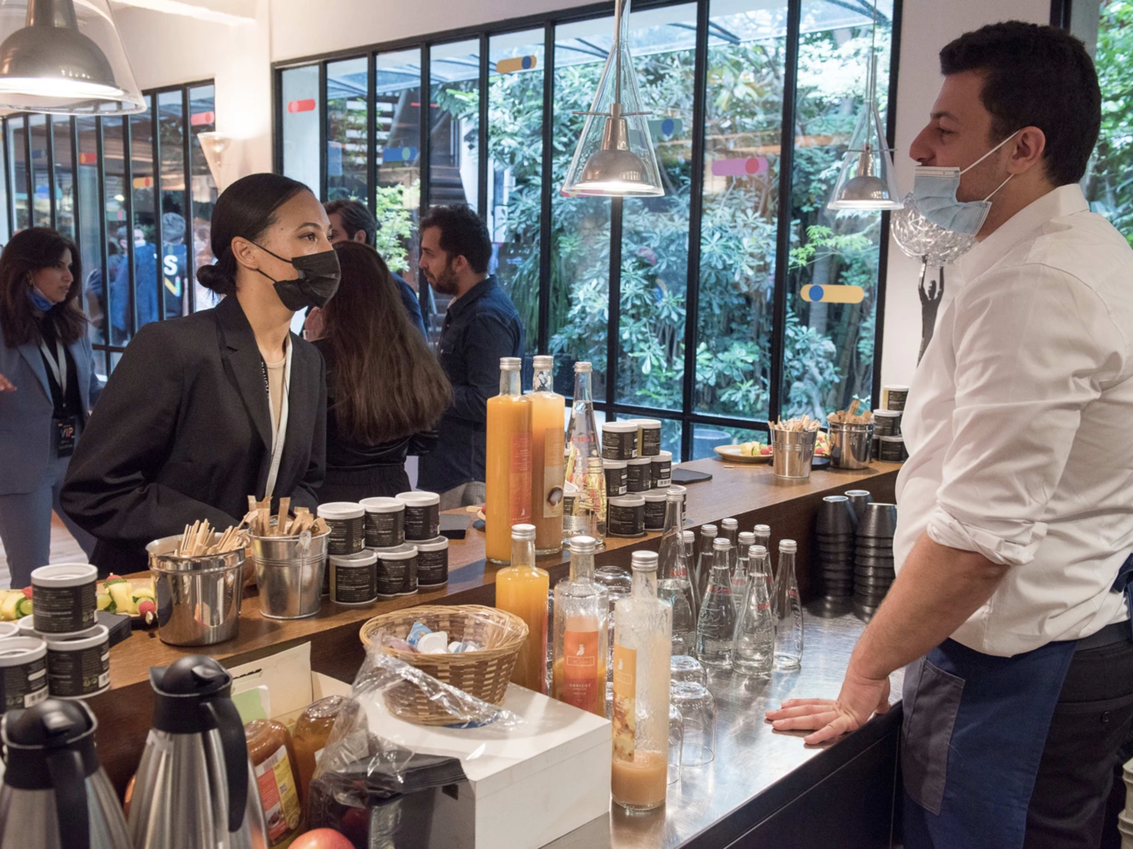 A woman in a black mask talks to a man in a white shirt at a coffee shop counter filled with beverages and snacks, while greenery is visible through large windows behind them.
