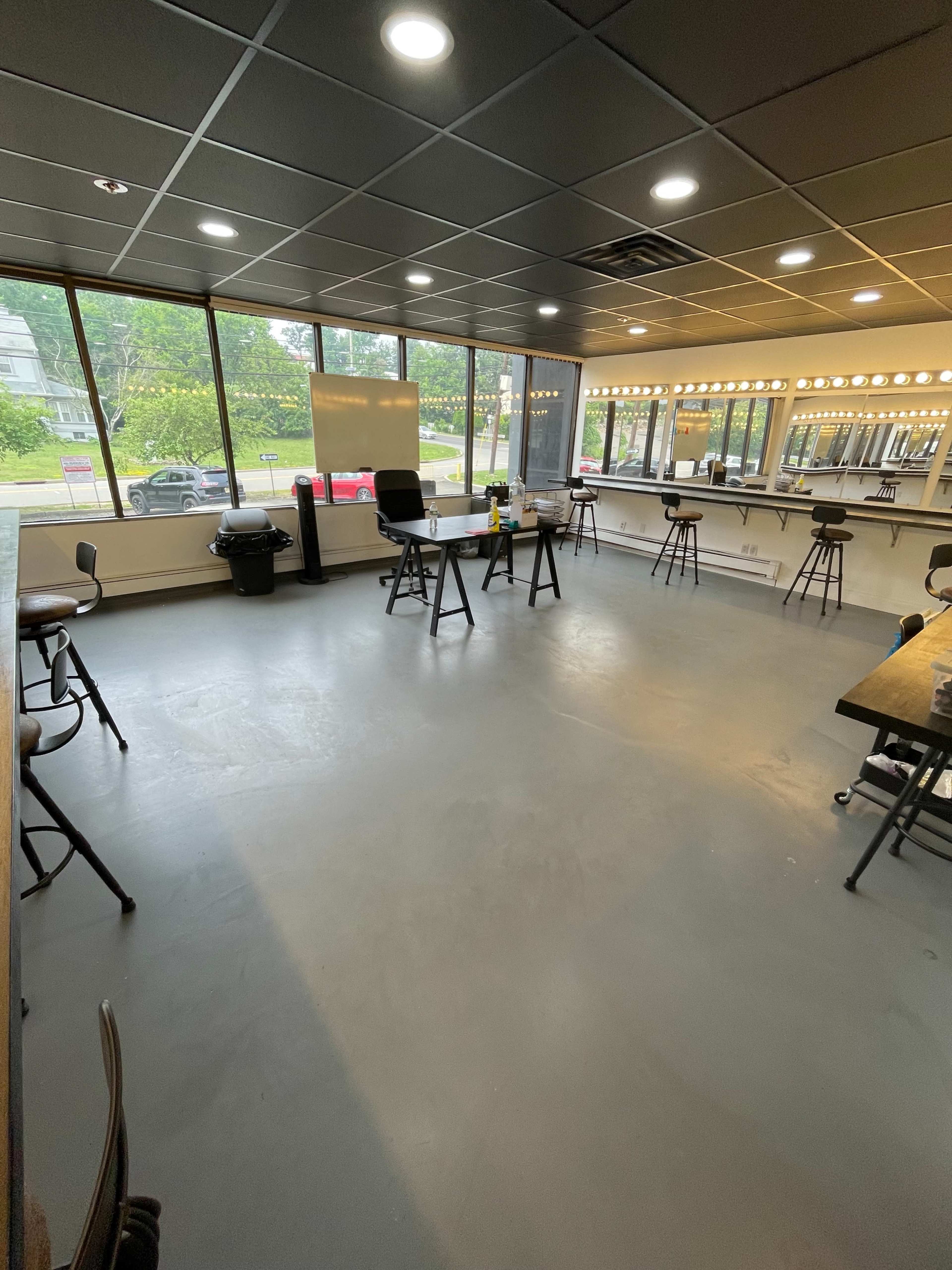 An empty room with large windows, a few tables, and bar stools, featuring a whiteboard and a trash can.