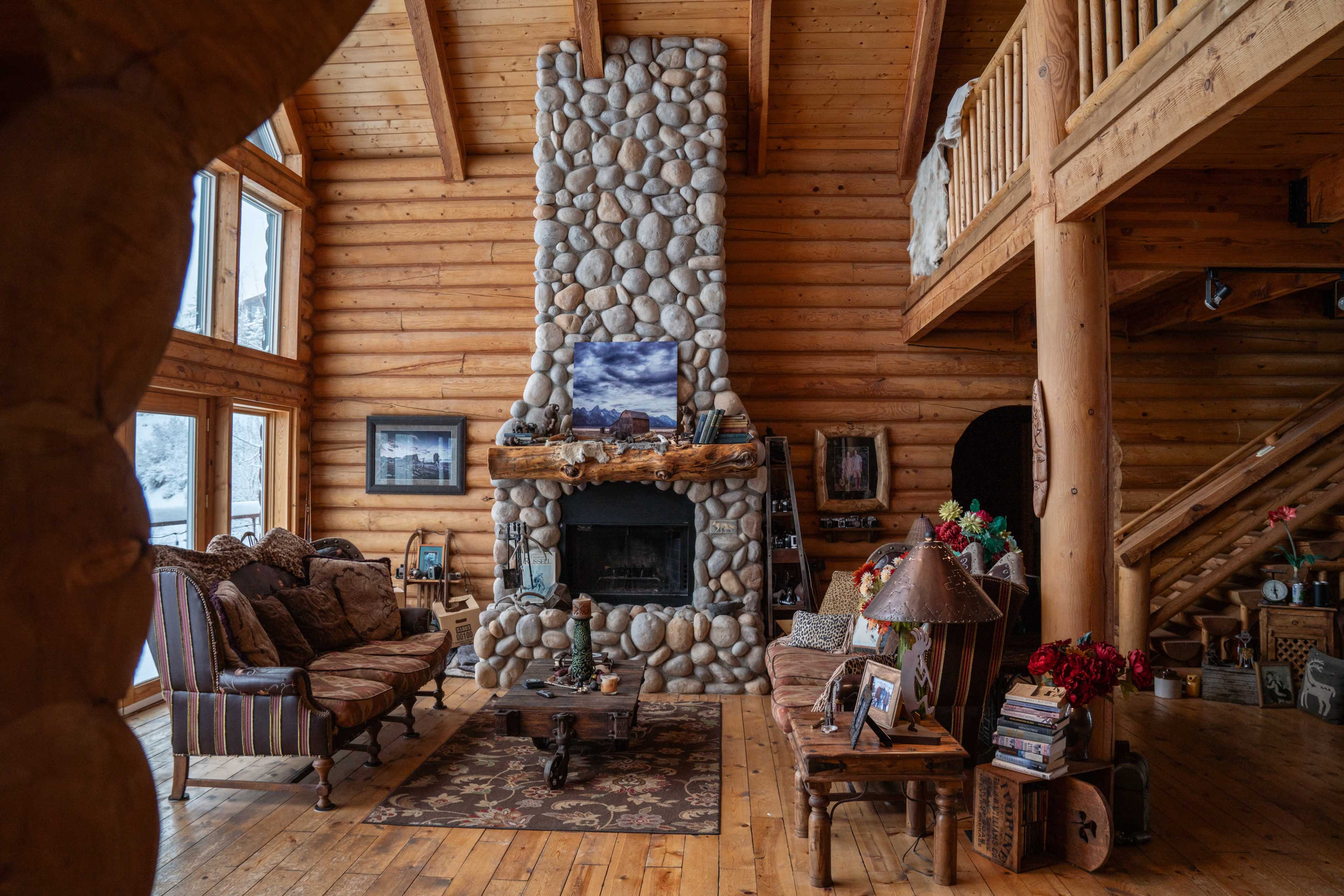 A rustic wooden interior of a cabin with a stone fireplace, large windows, and a cozy seating area.