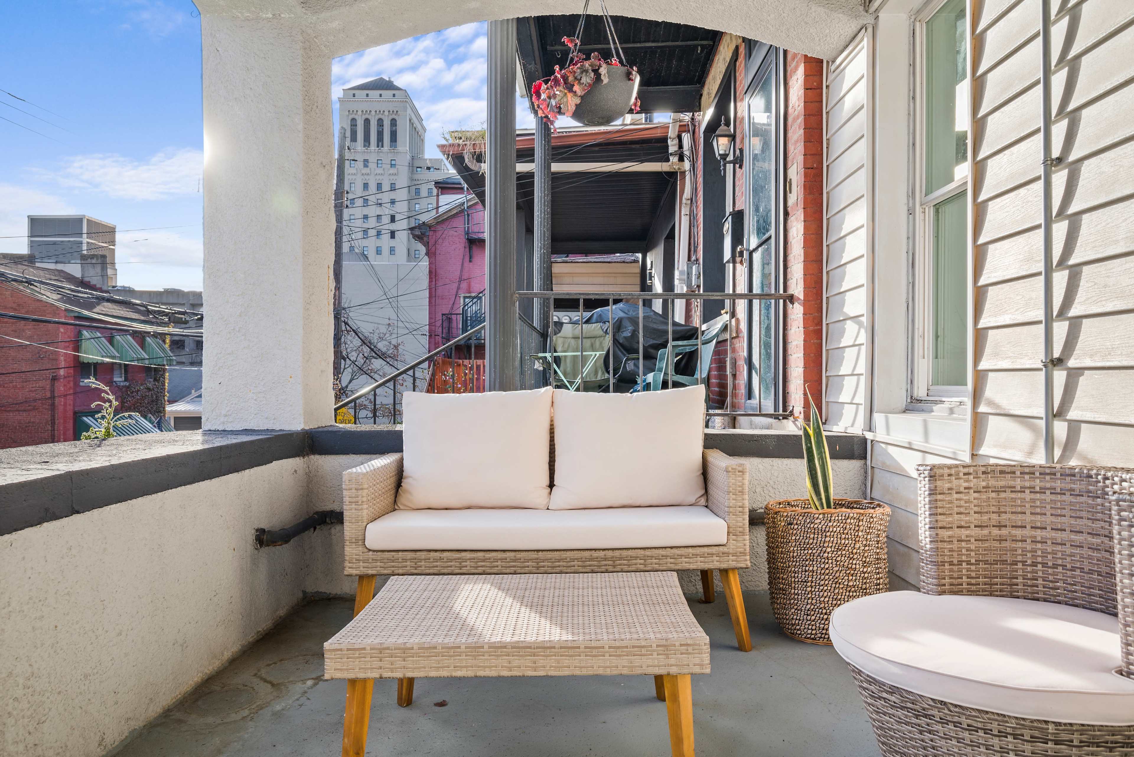 A small balcony furnished with a wicker couch and a table, overlooking a cityscape with buildings in the background.