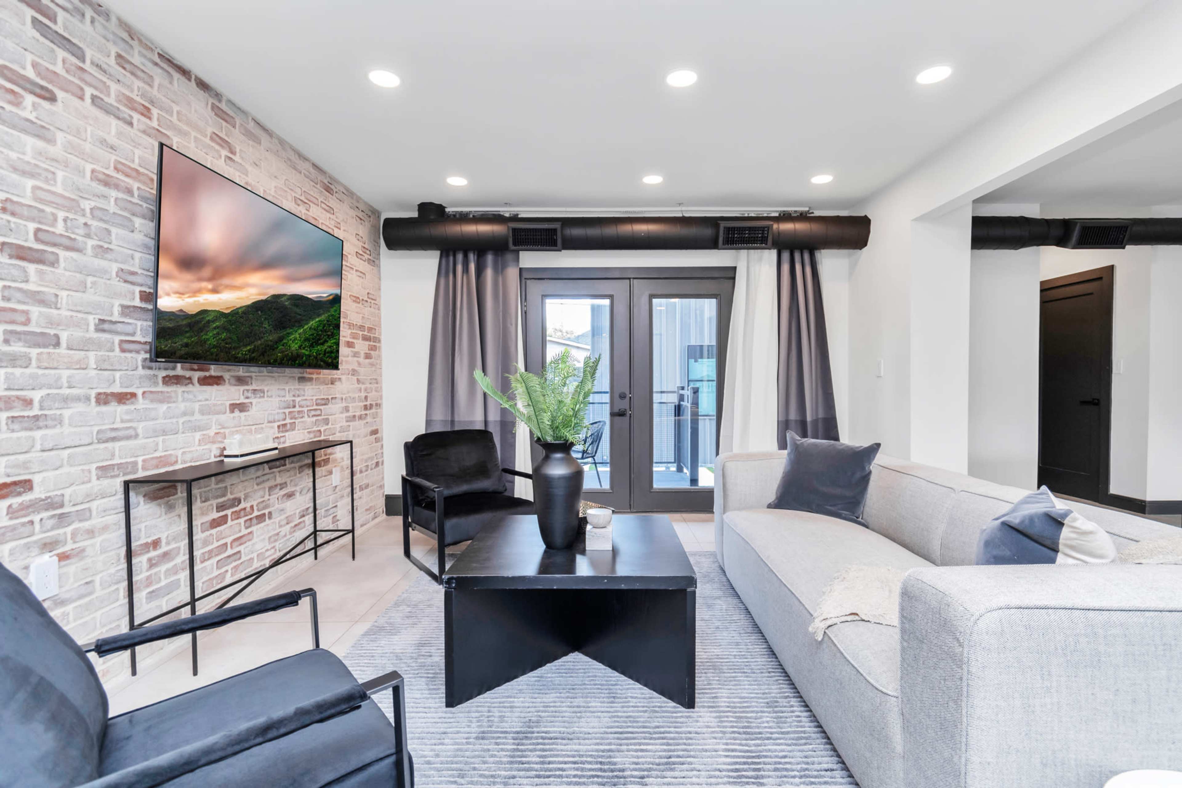 A modern living room with a gray sofa, two black chairs, a coffee table, and a large brick wall with a mounted television.