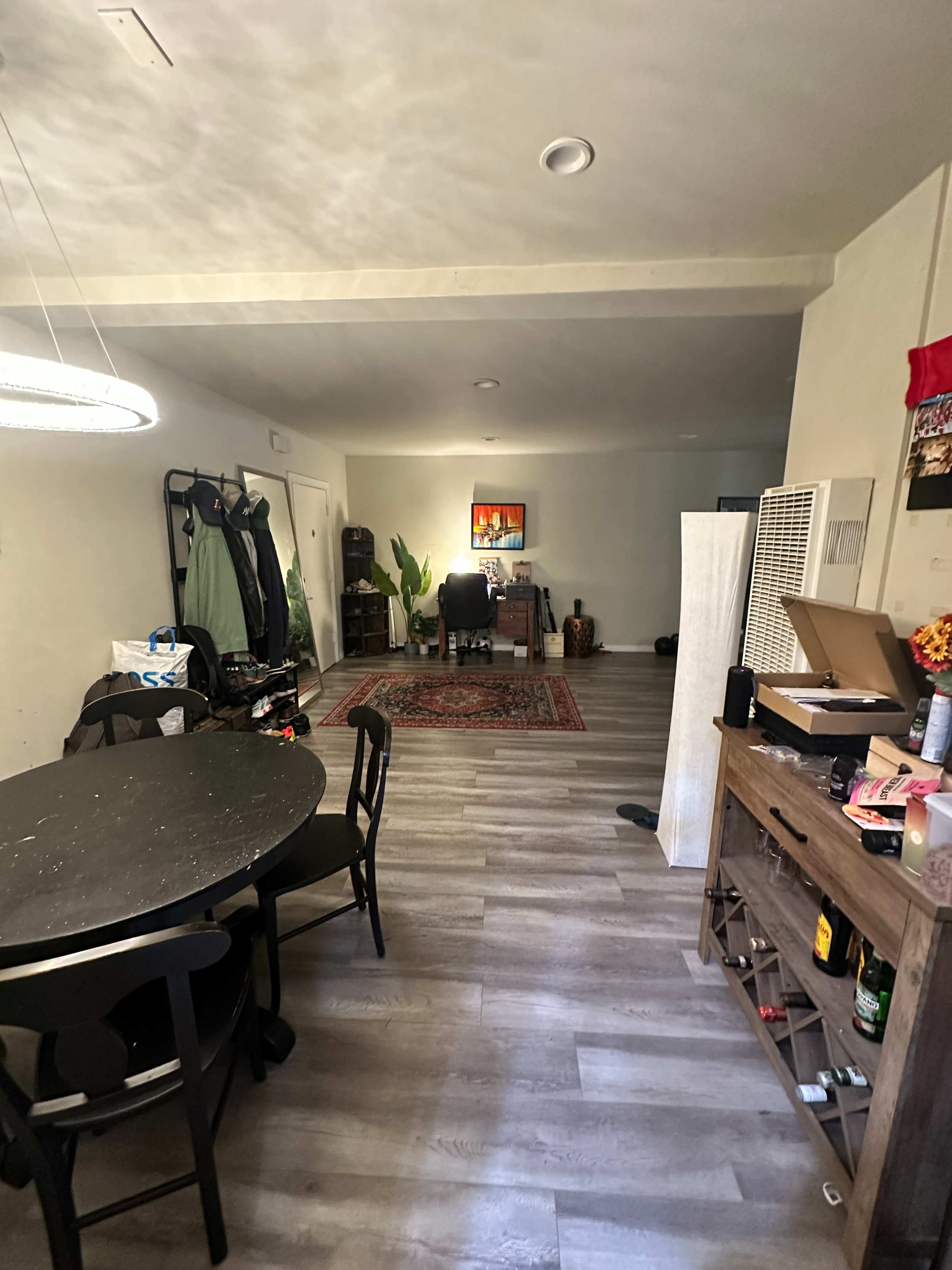 The image shows a spacious room with a dining area featuring a round black table, a light fixture above, and a view of a living area with a desk and plants in the background.