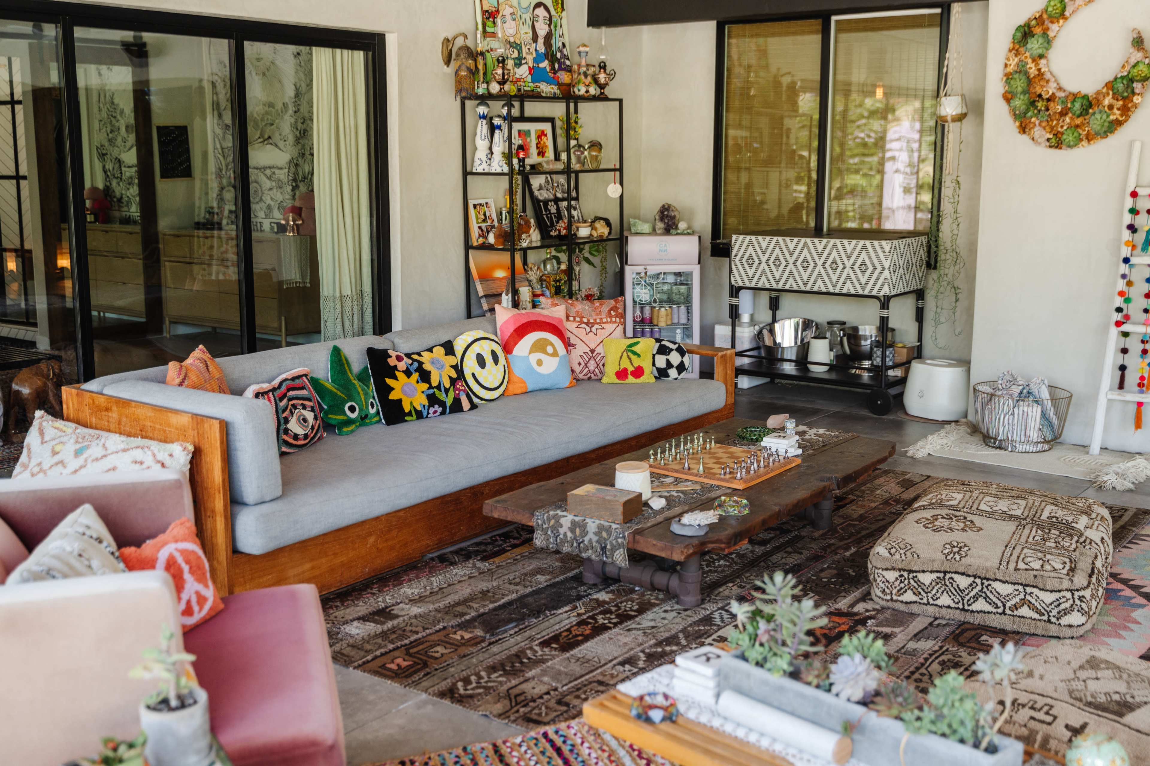 A cozy living room with a gray sofa adorned with colorful decorative pillows, a wooden coffee table, and various shelves displaying decorative items and plants.