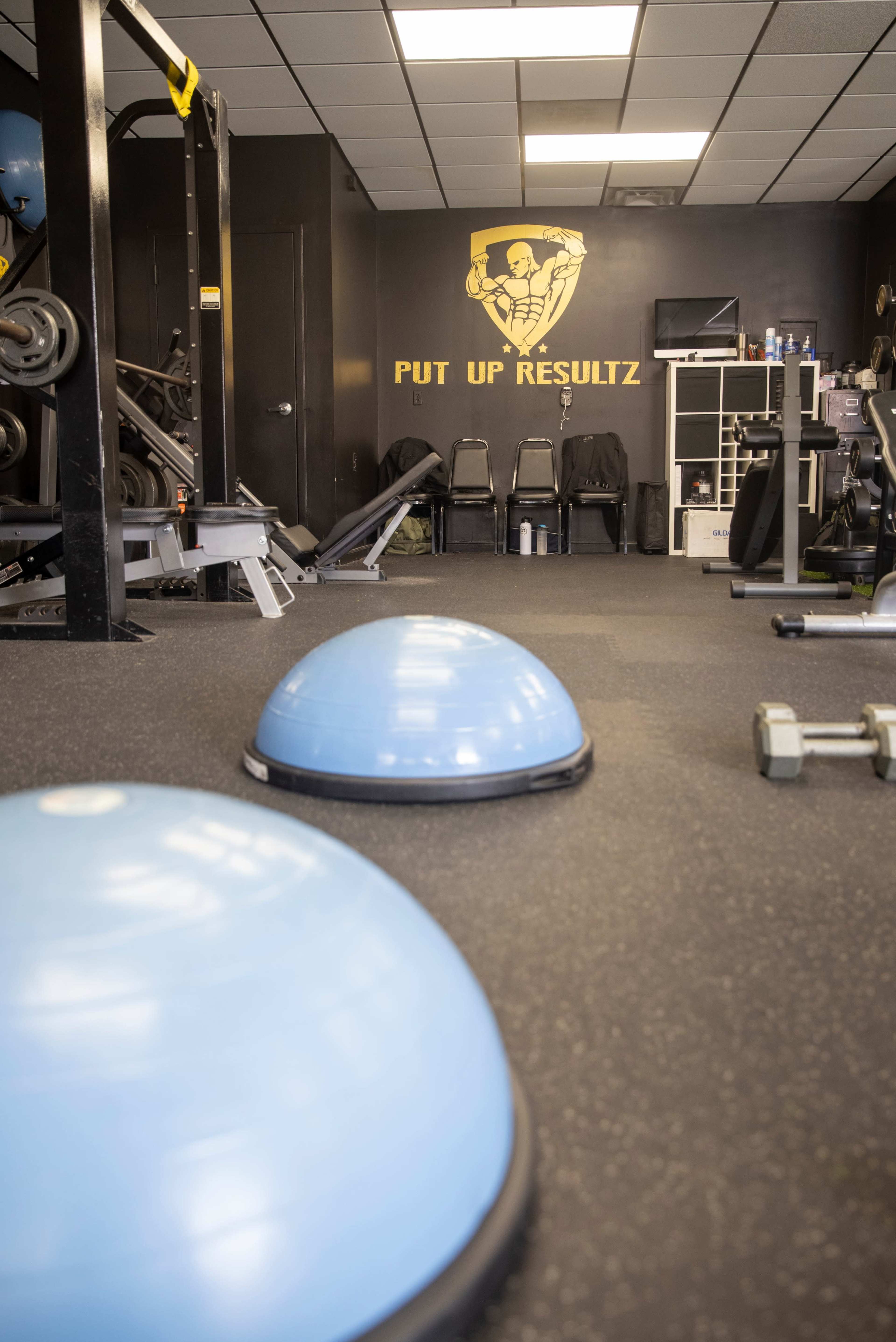 A fitness gym with blue balance trainers on the floor and exercise equipment in the background, featuring a wall with the phrase "PUT UP RESULTZ."
