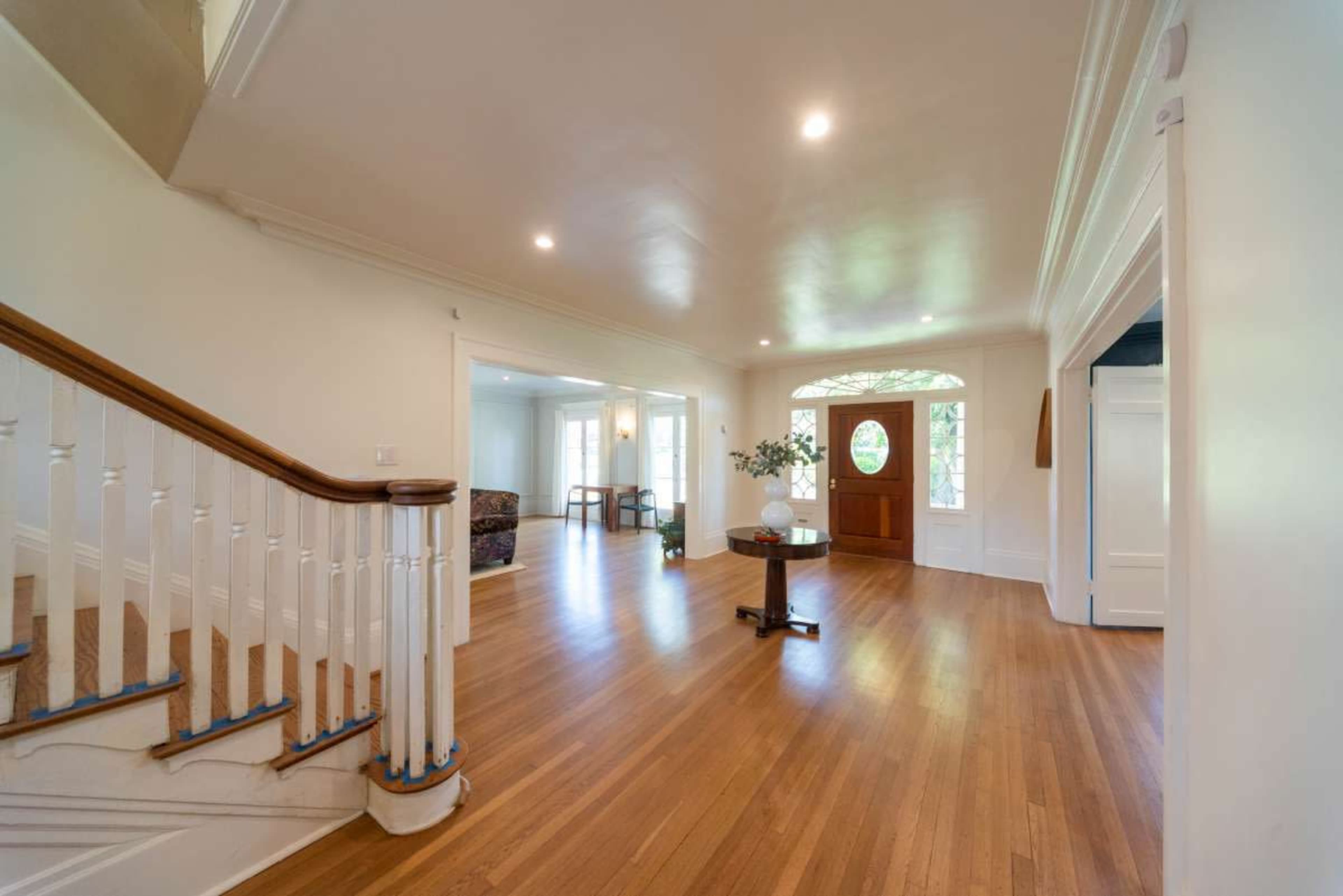 A spacious entryway features a wooden staircase, large windows, and a round table with a vase of flowers.
