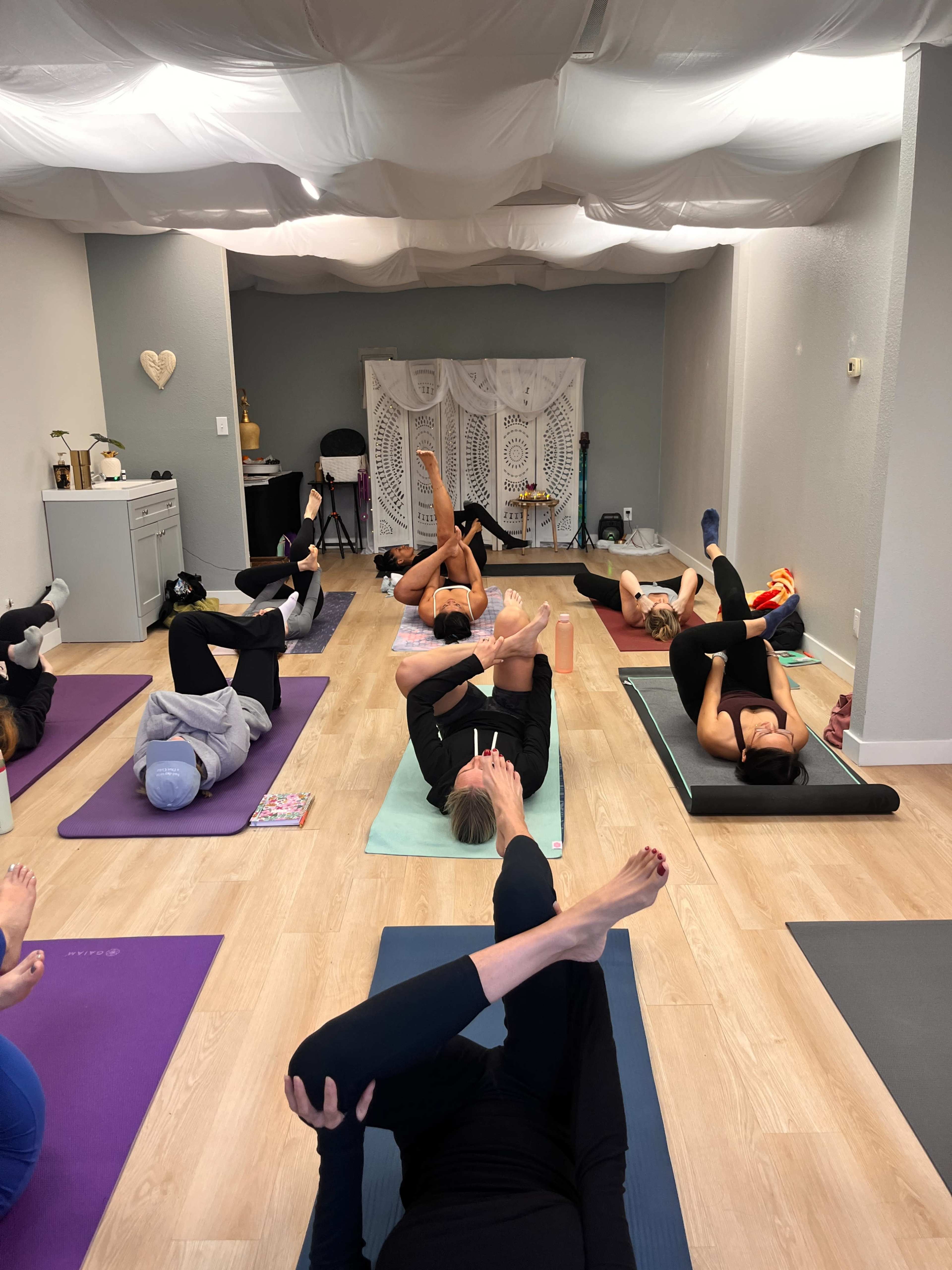 A group of people is participating in a yoga class on mats in a well-lit room with a white draped backdrop.