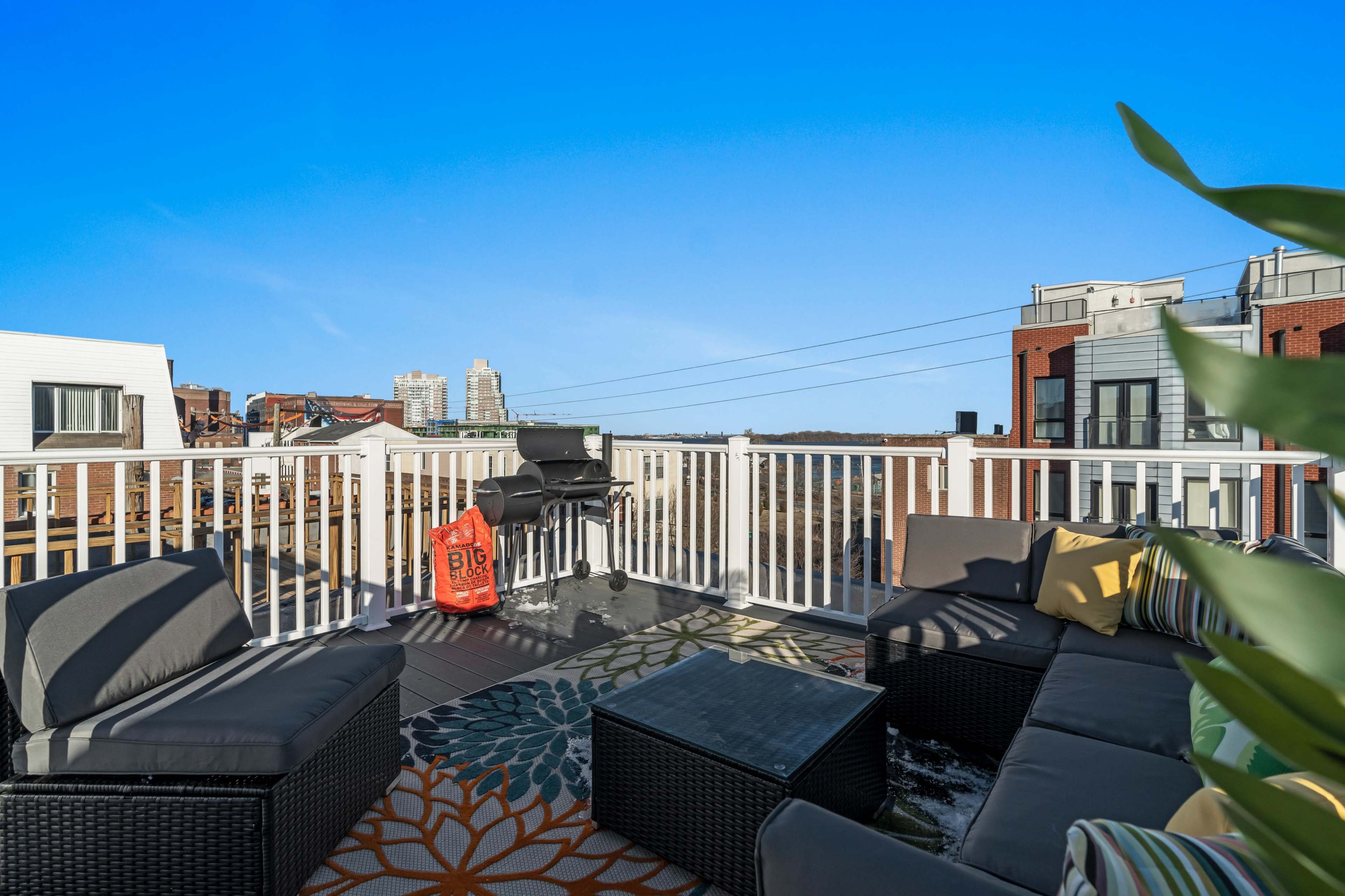 The image shows a rooftop terrace with seating areas, a barbecue grill, and a clear blue sky in the background.
