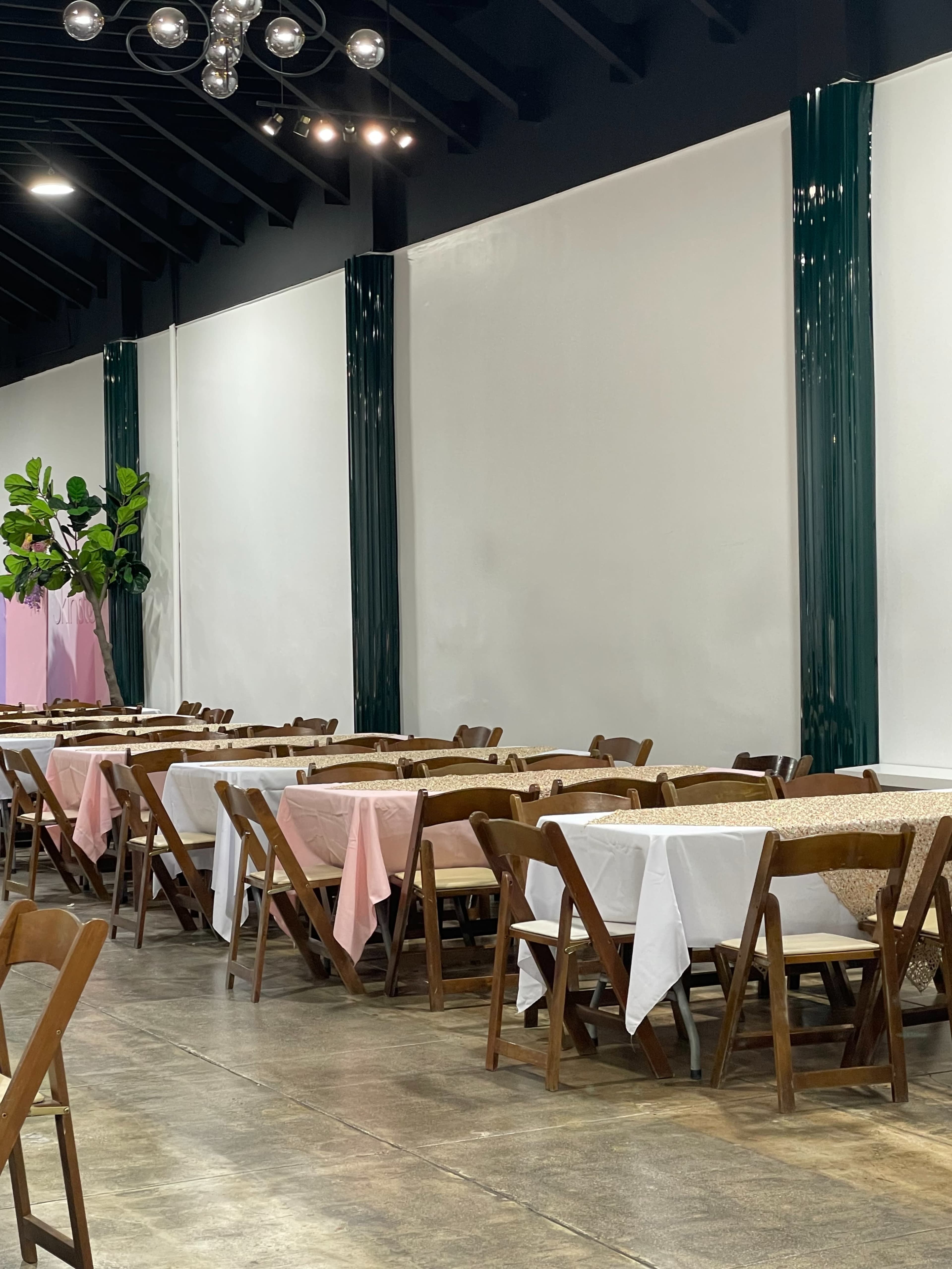 The image shows a dining area with wooden folding chairs and tables covered in pink, white, and gold tablecloths, set against a plain wall with decorative greenery in the background.