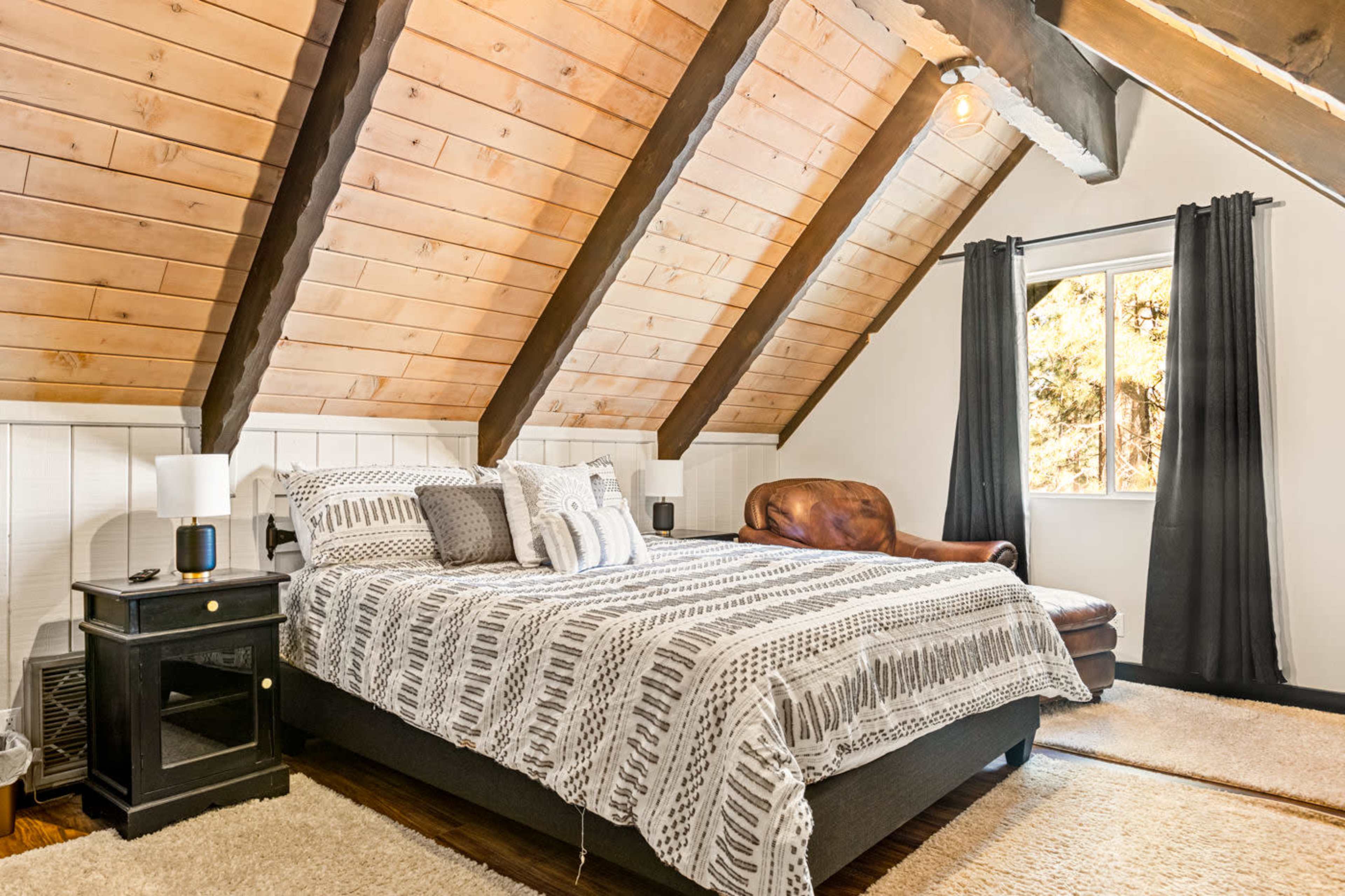 The bedroom features an A-frame design with wooden beams, a bed with patterned bedding, a brown leather chair, and a window with dark curtains.