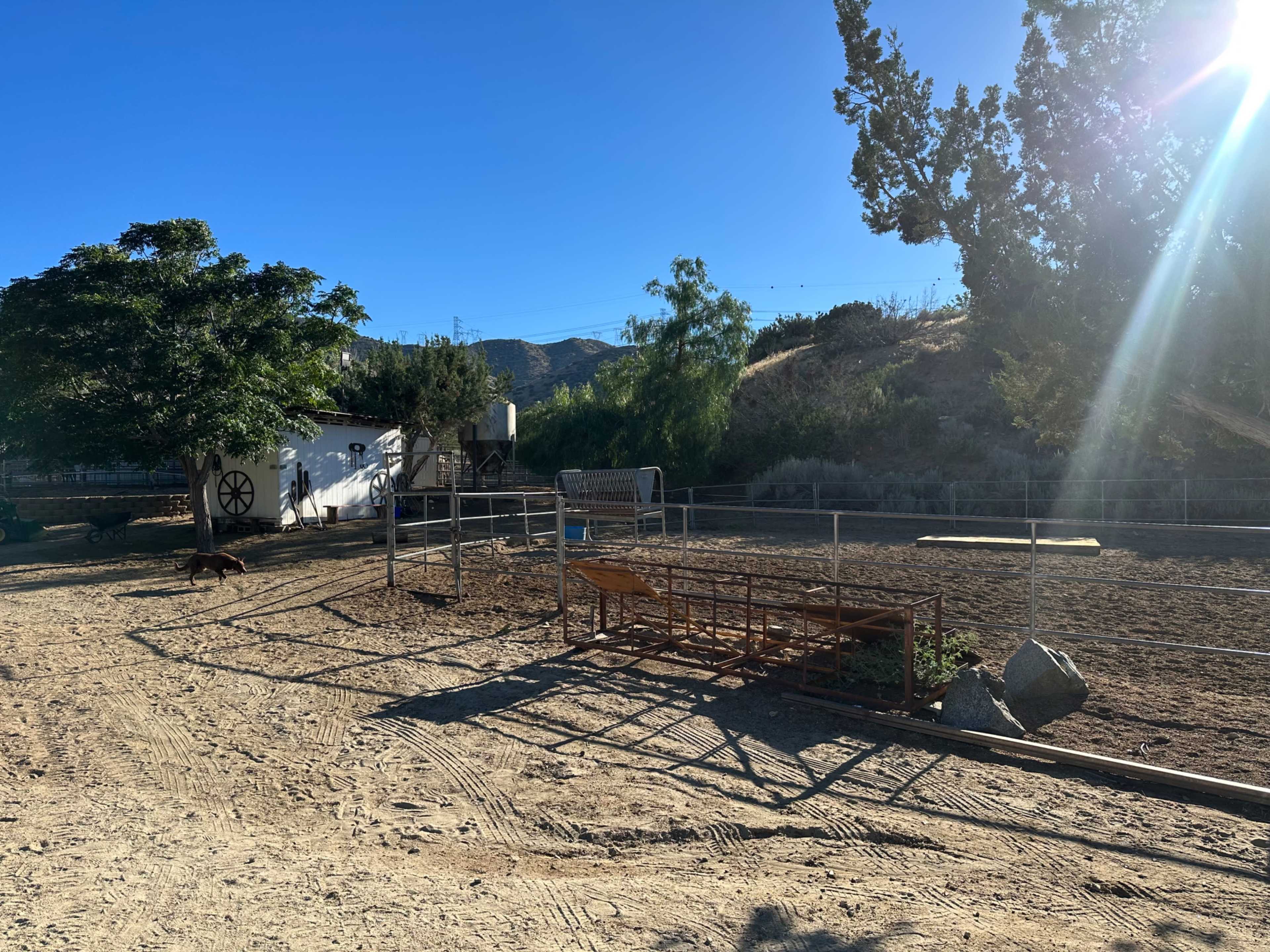 A fenced area on a ranch with dirt ground, a wooden structure, and distant hills under a clear blue sky.