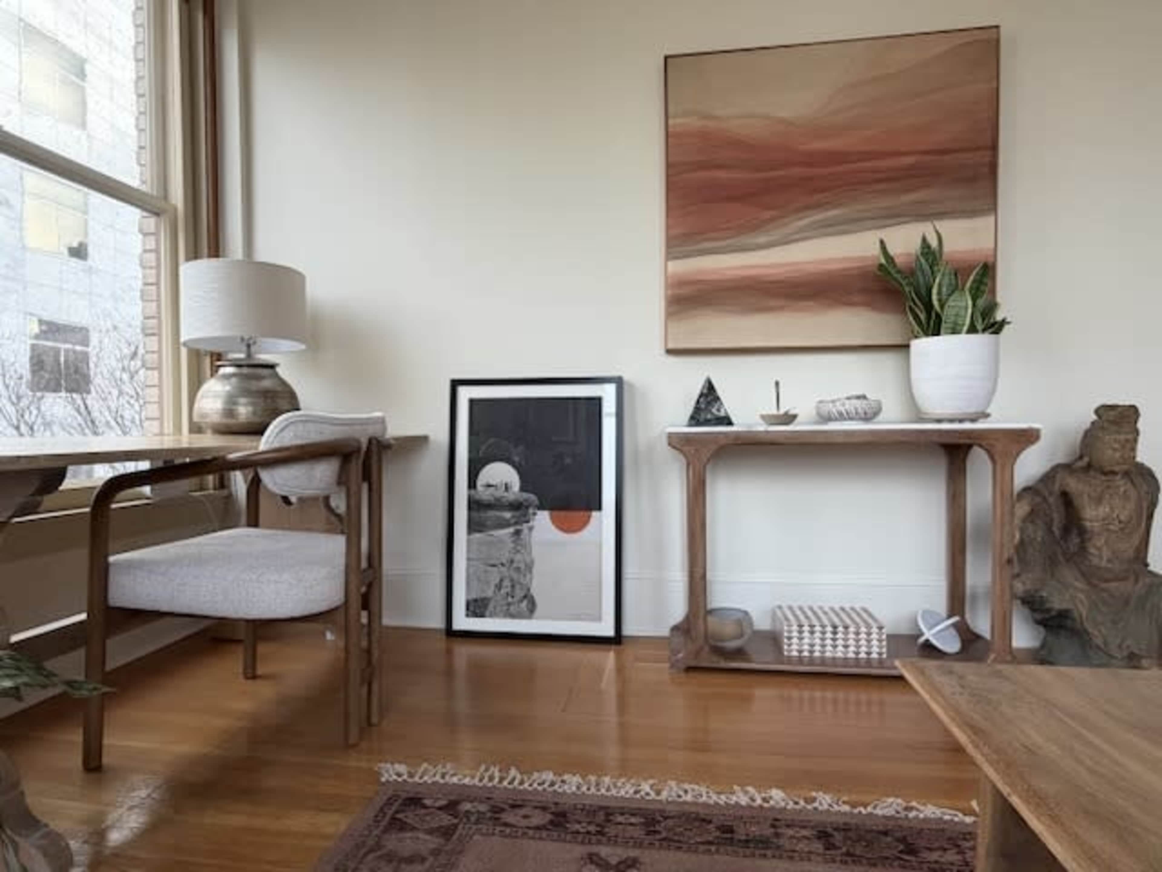 The image shows a cozy interior setting featuring a wooden table, a framed artwork, a lamp, and decorative plants, with a large abstract painting on the wall.
