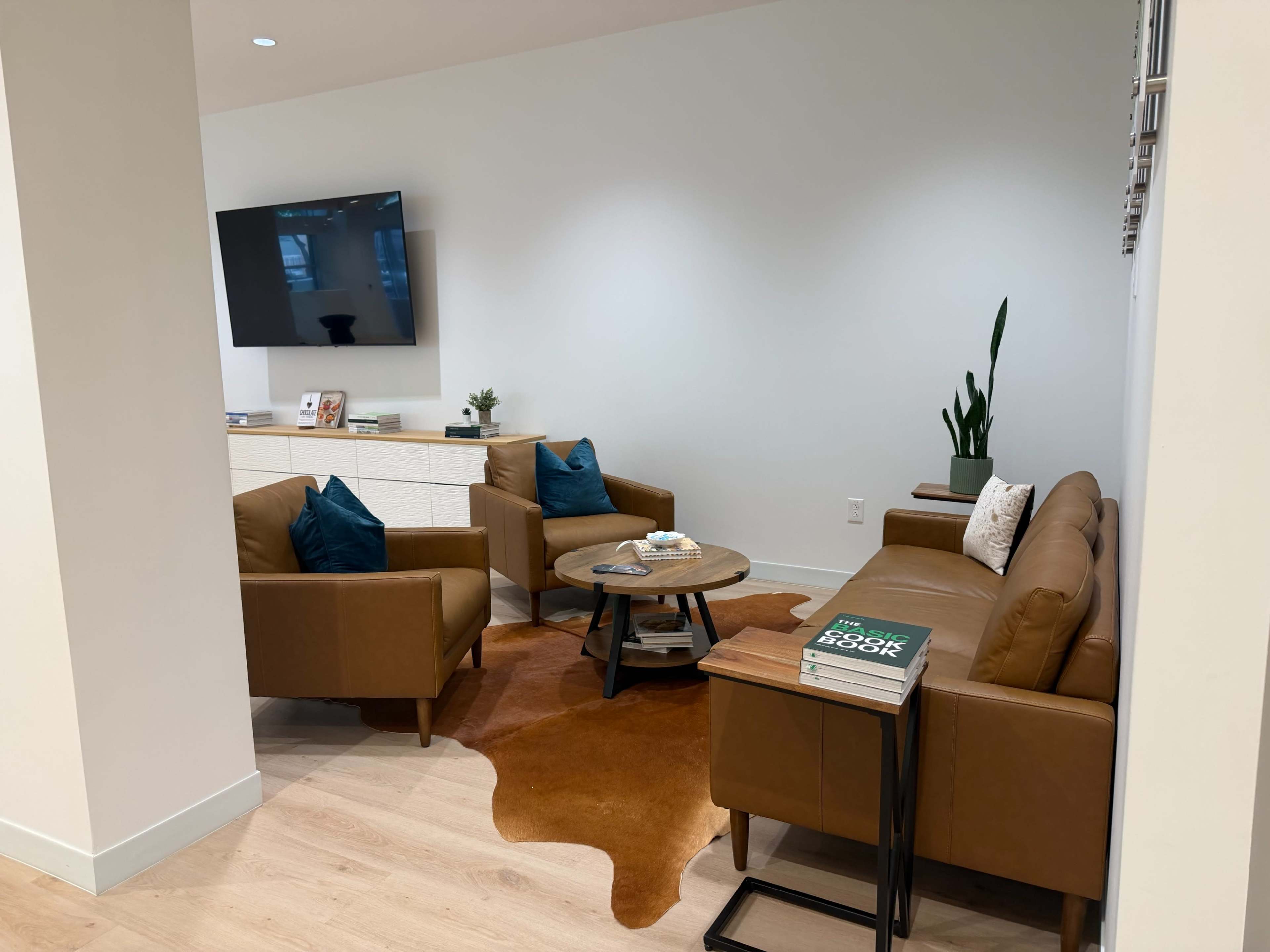 The image shows a modern living room featuring two brown leather chairs and a sofa, a small coffee table, a decorative plant, and a wall-mounted television.