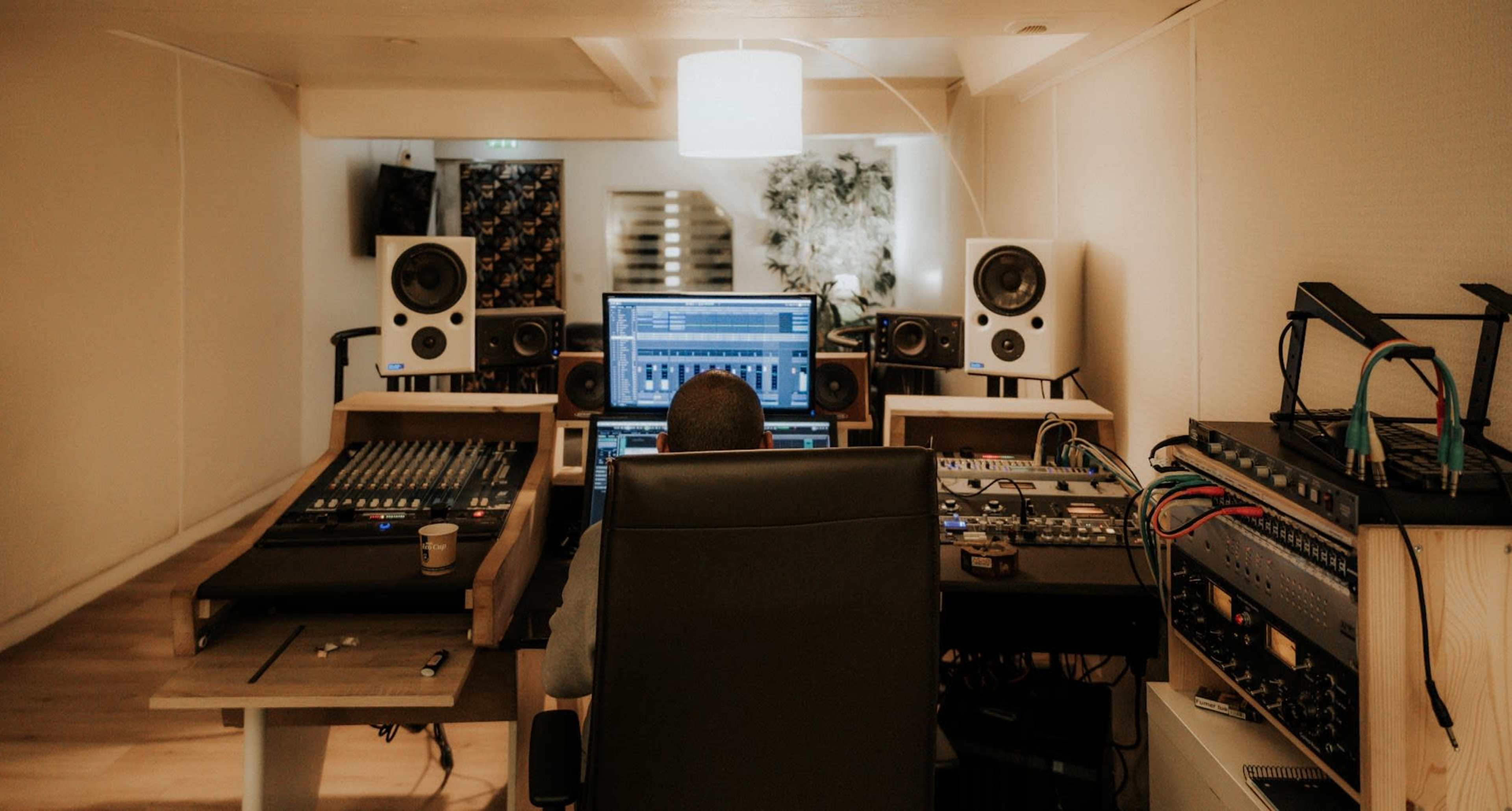 A person sits in a recording studio surrounded by audio equipment, monitors, and mixing consoles while working on a computer.