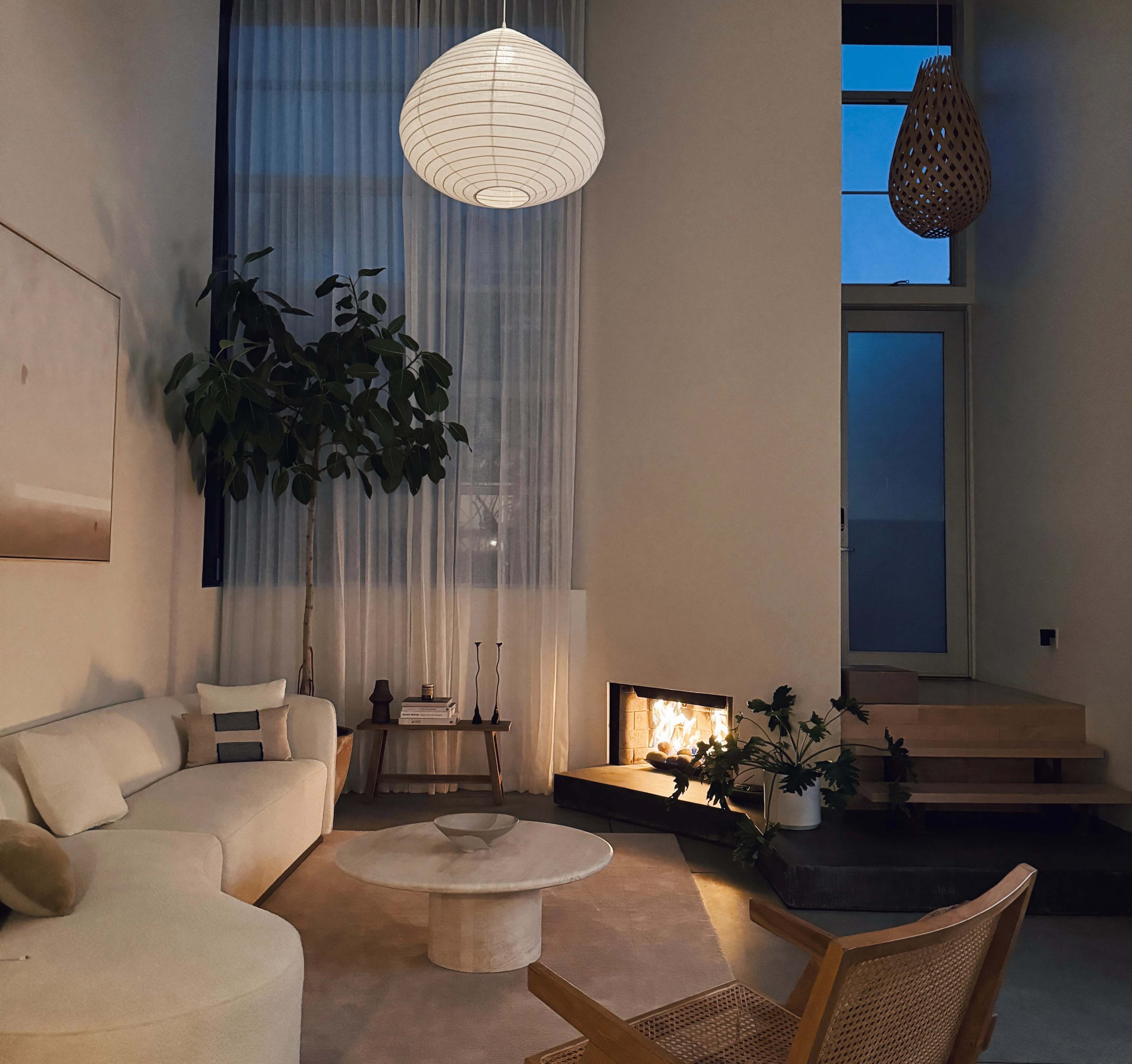 The image shows a modern living room featuring a large sofa, a round coffee table, a fireplace, and large windows with sheer curtains, illuminated by soft lighting.