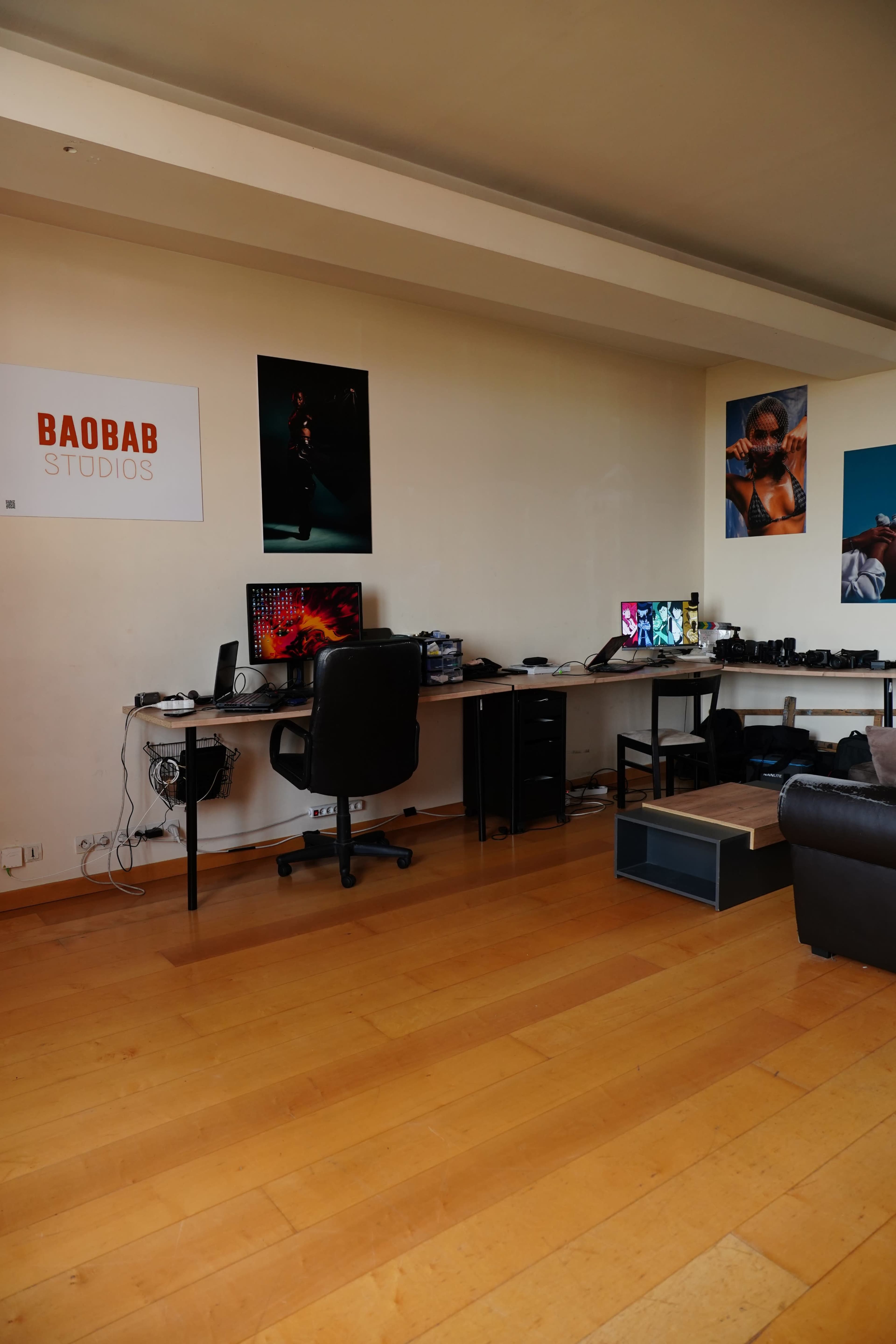 A studio workspace with two desks, computers, and various equipment against a wall adorned with posters.