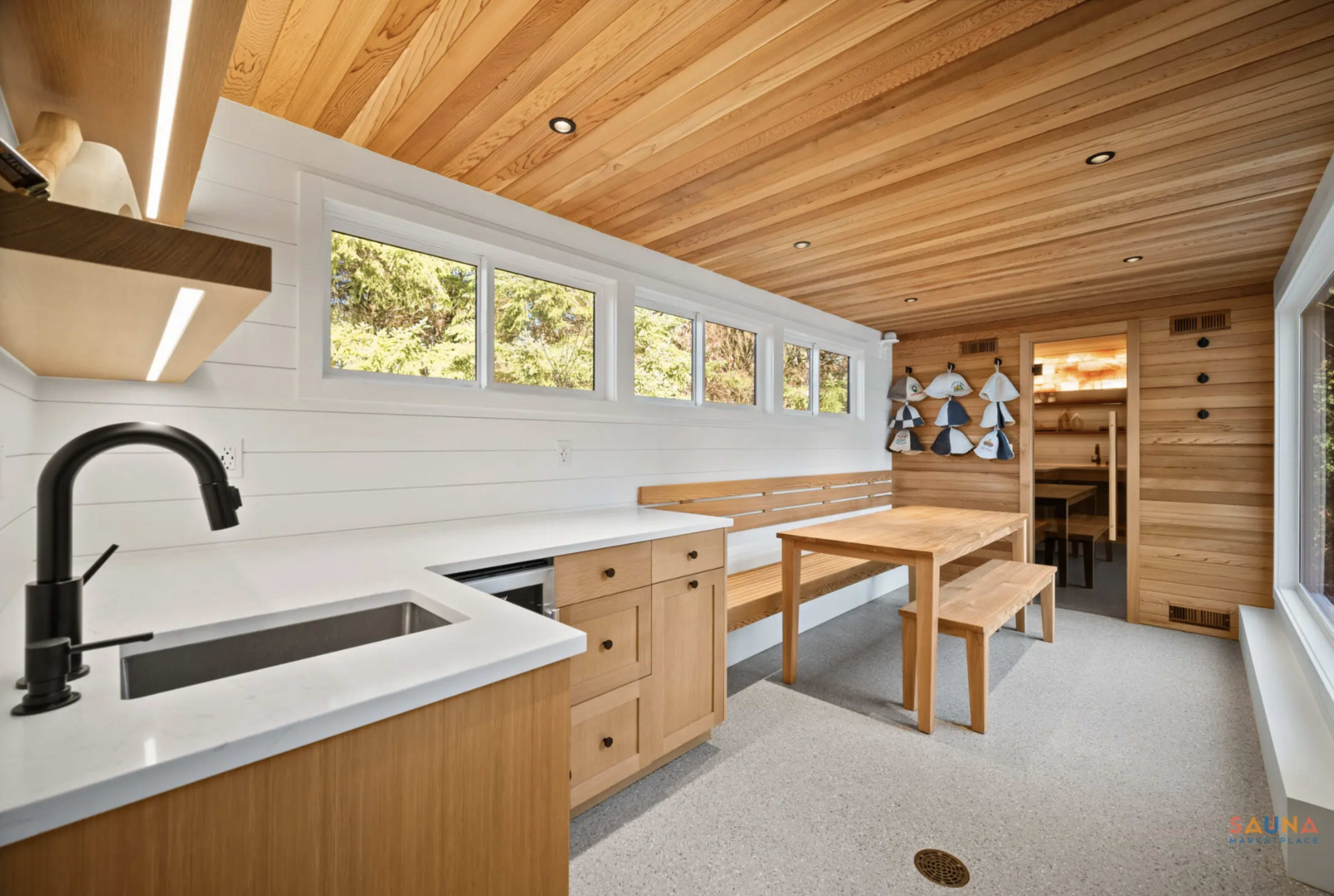 The image shows a modern kitchen and dining area featuring wooden ceilings, a large wooden table, and a minimalist design with a black faucet and white countertops.