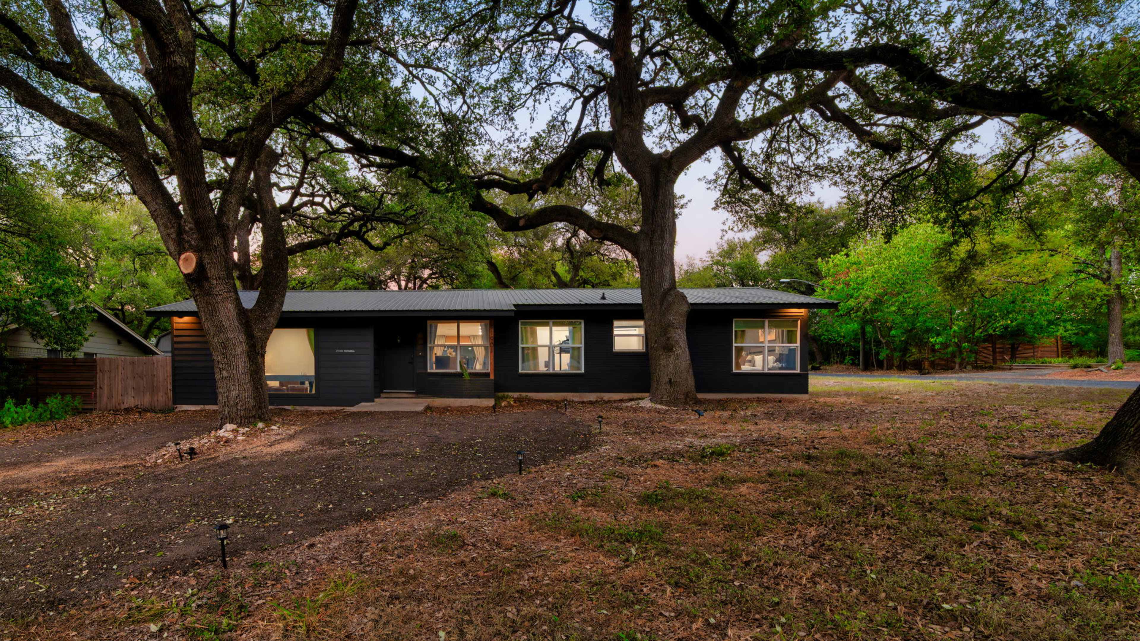 A single-story black house is surrounded by oak trees and features large windows.