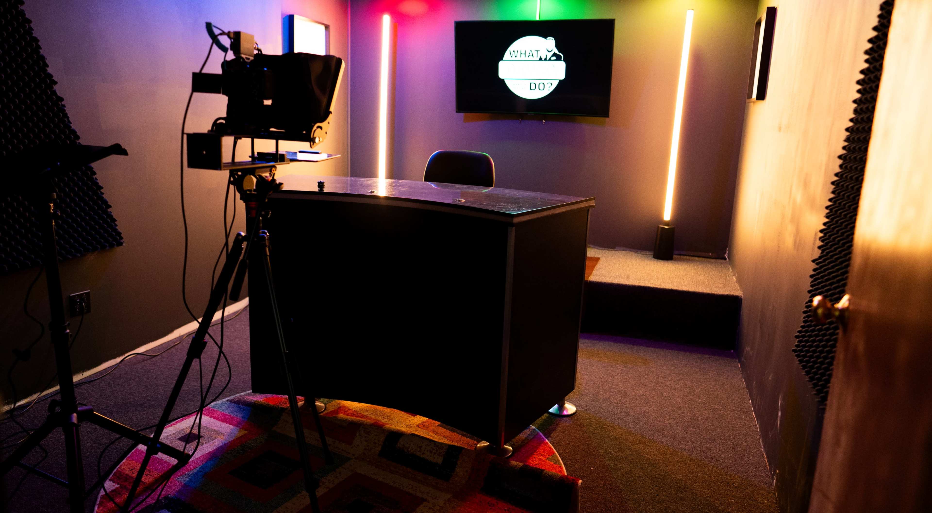 A desk and chair are set up in a small room equipped with colorful lights and a television screen displaying a logo.