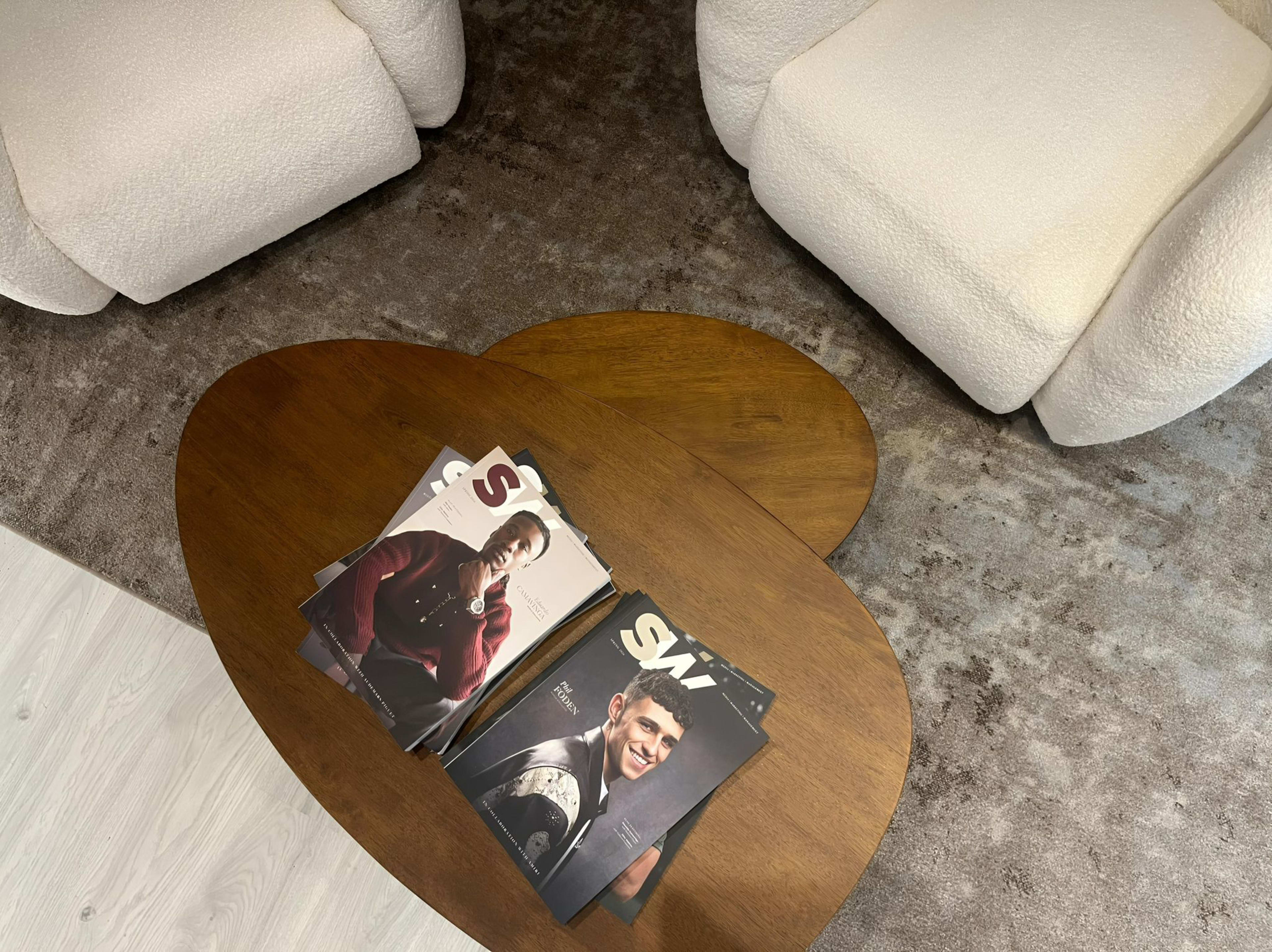 A wooden coffee table features a stack of magazines on a textured rug with two upholstered armchairs nearby.