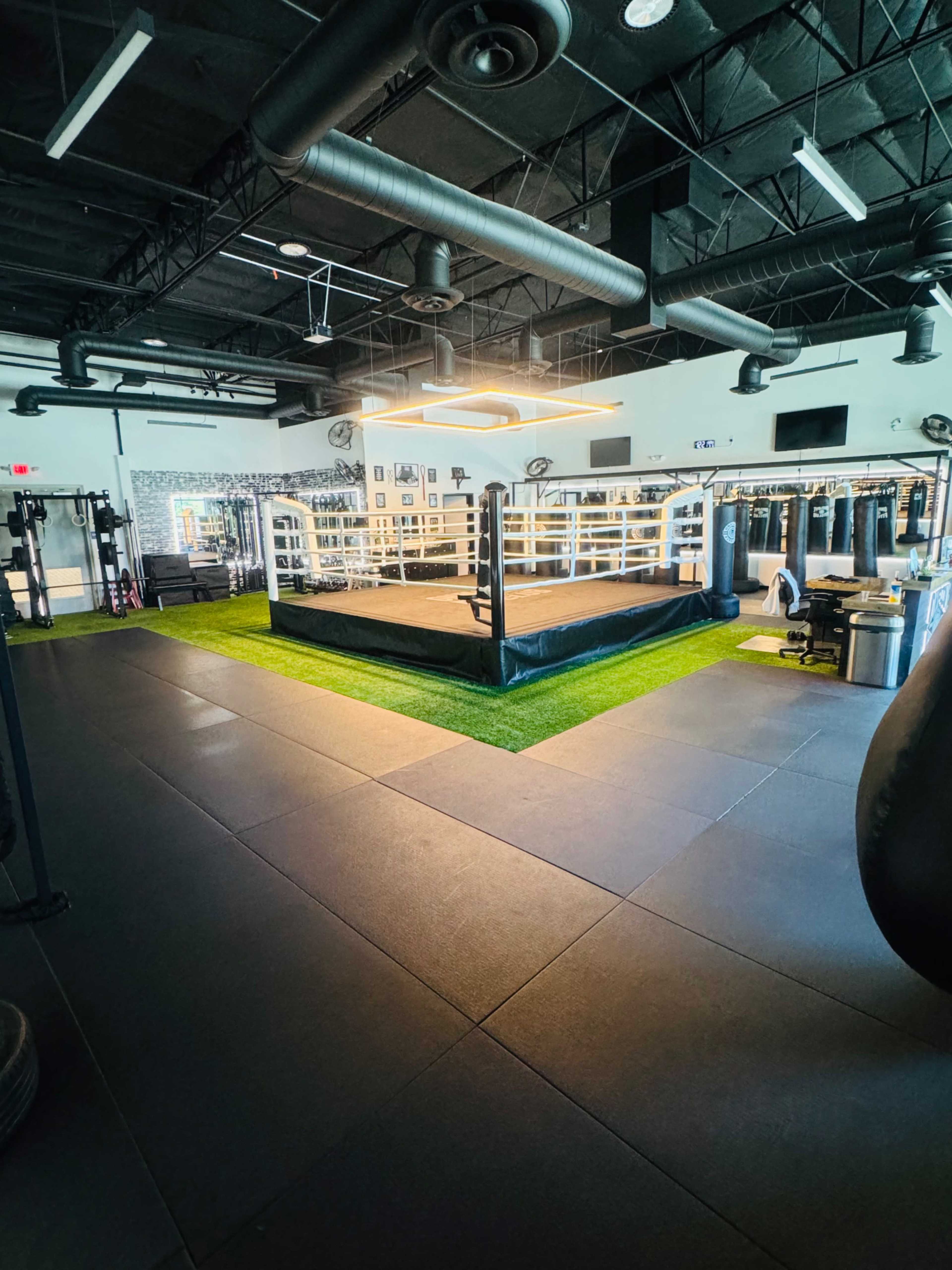 A spacious boxing gym featuring a central ring surrounded by exercise equipment and padded flooring.