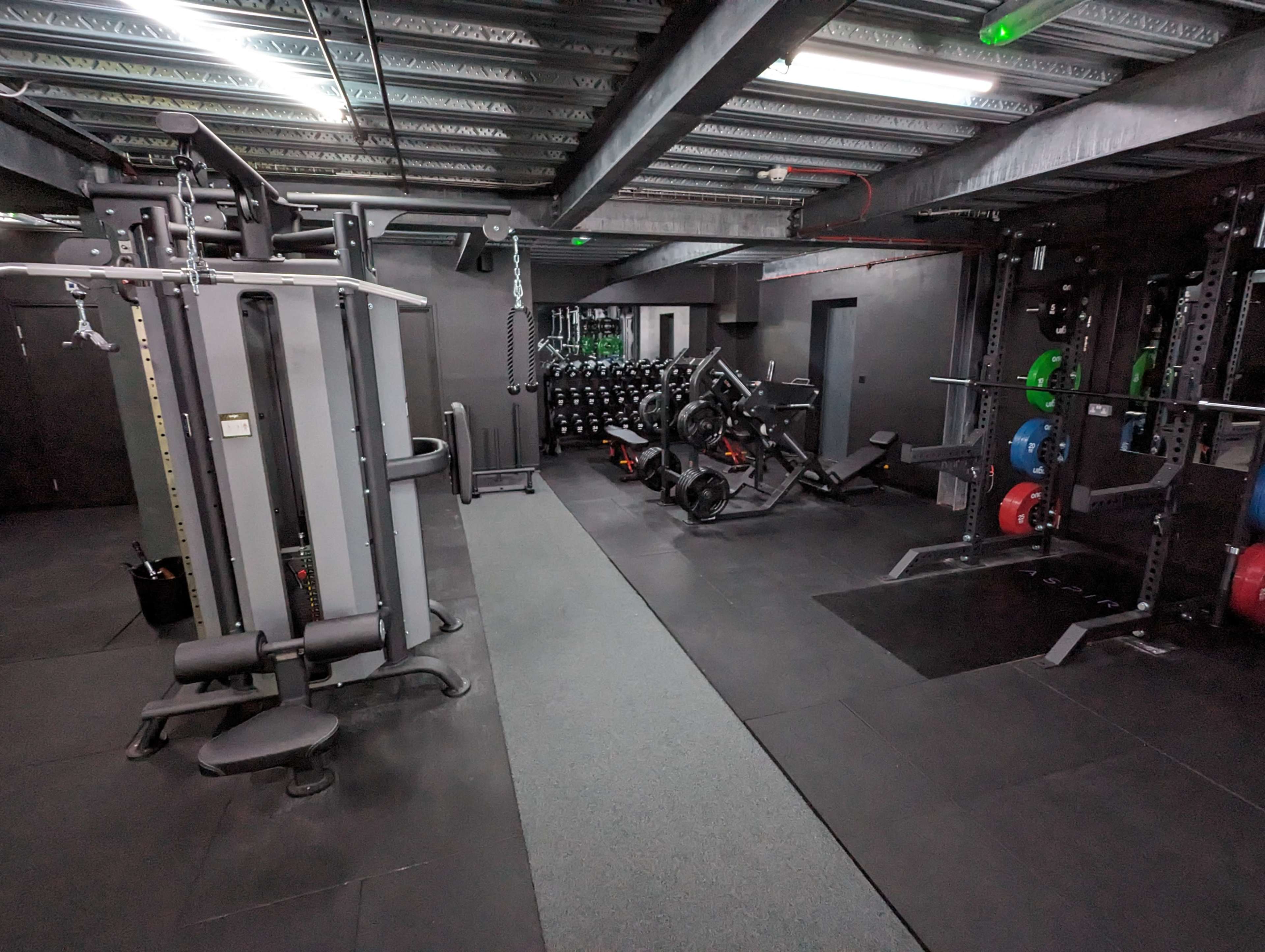 A spacious gym equipped with various weightlifting machines, free weights, and exercise mats on a black rubber floor.