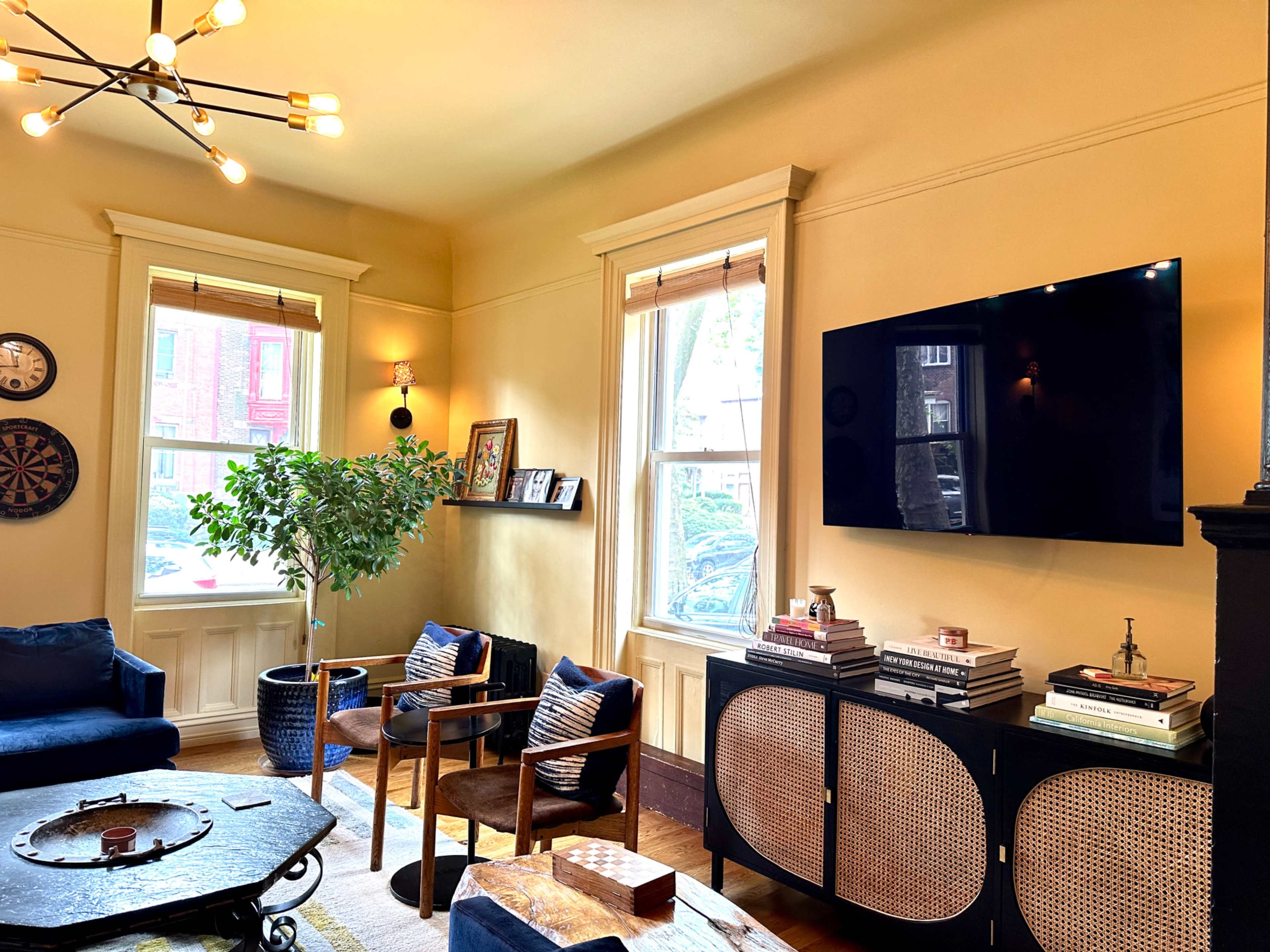 A cozy living room featuring a television mounted on a wall, a dark cabinet with decorative books, a potted plant, and a seating area with blue chairs and a circular table.
