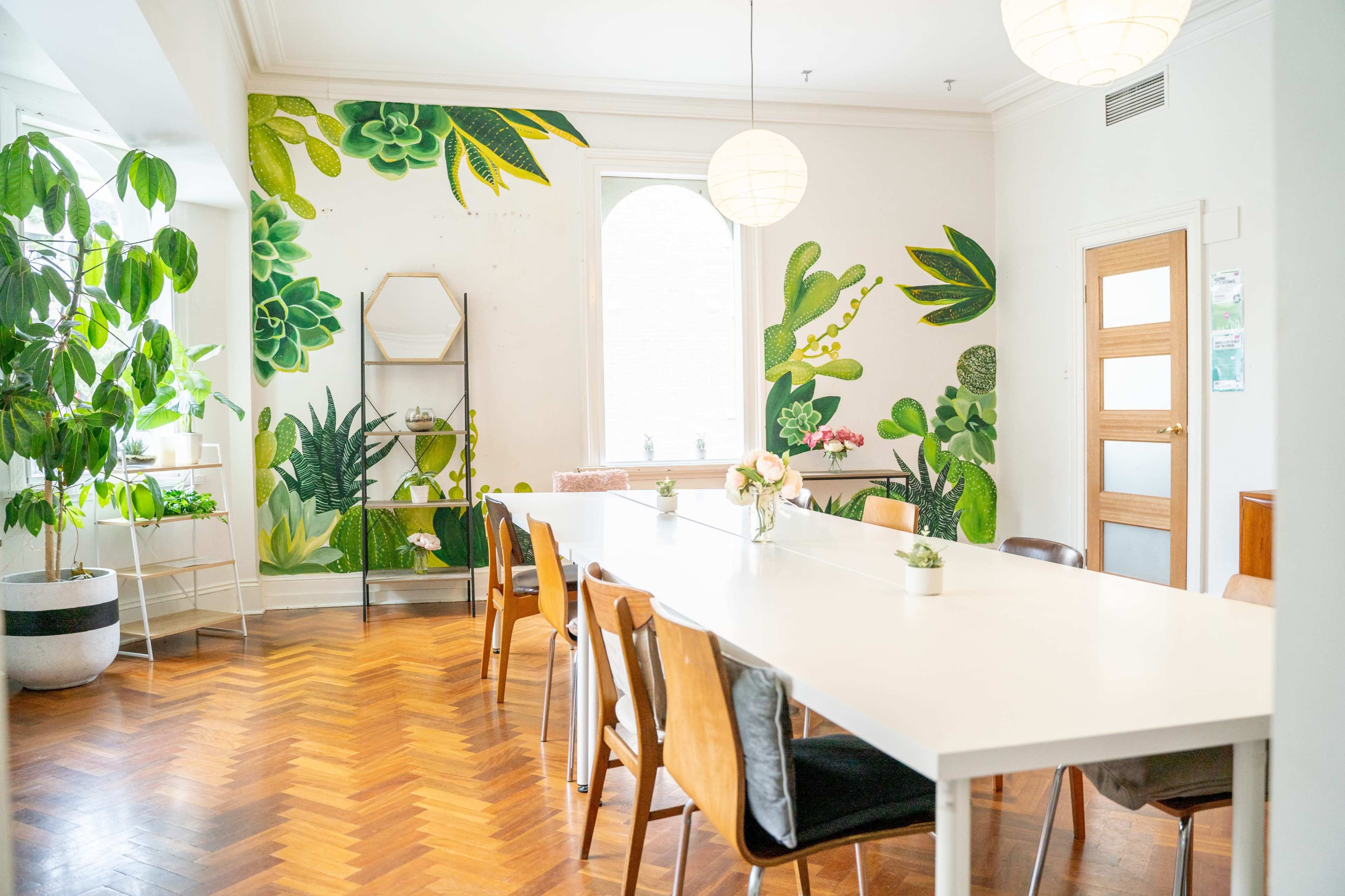 A bright dining area featuring a large white table surrounded by wooden chairs, with green plants and a botanical mural on the wall.