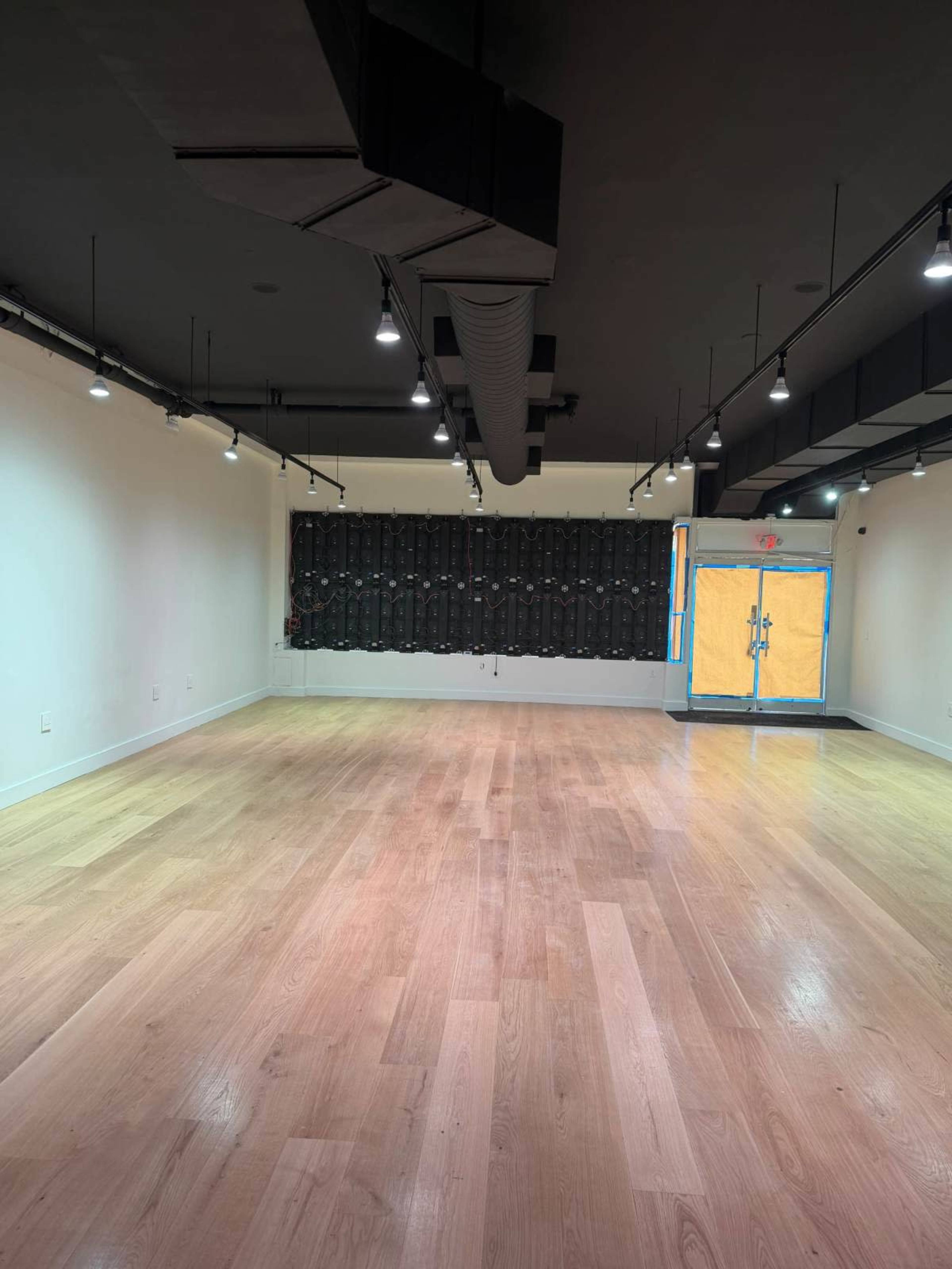 The image shows a spacious, empty room with wooden flooring, dimmable lights, and a black wall with a series of panels.