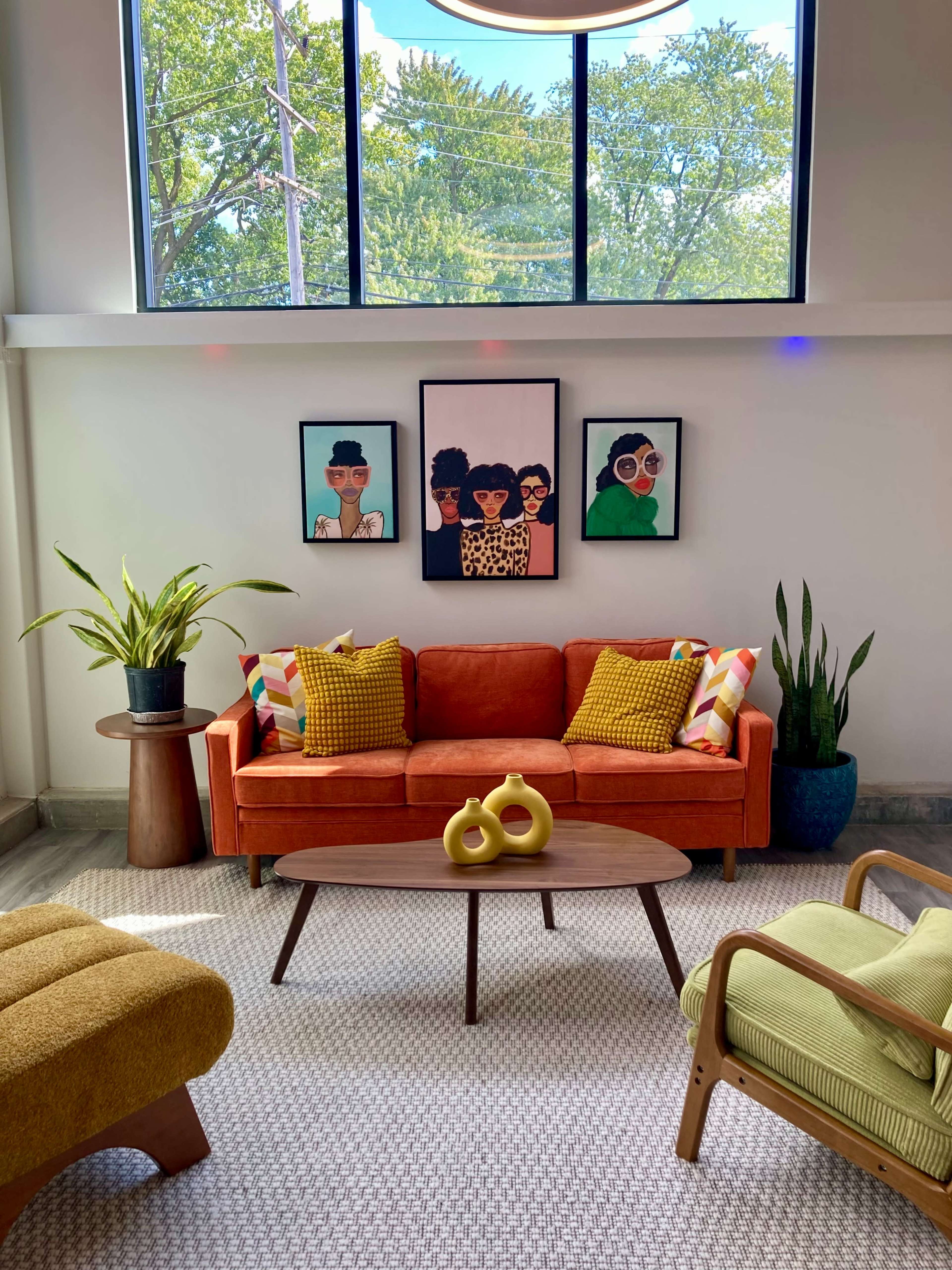 The scene features a vibrant living room with an orange sofa adorned with colorful cushions, flanked by potted plants, and decorated with three framed portraits on the wall.