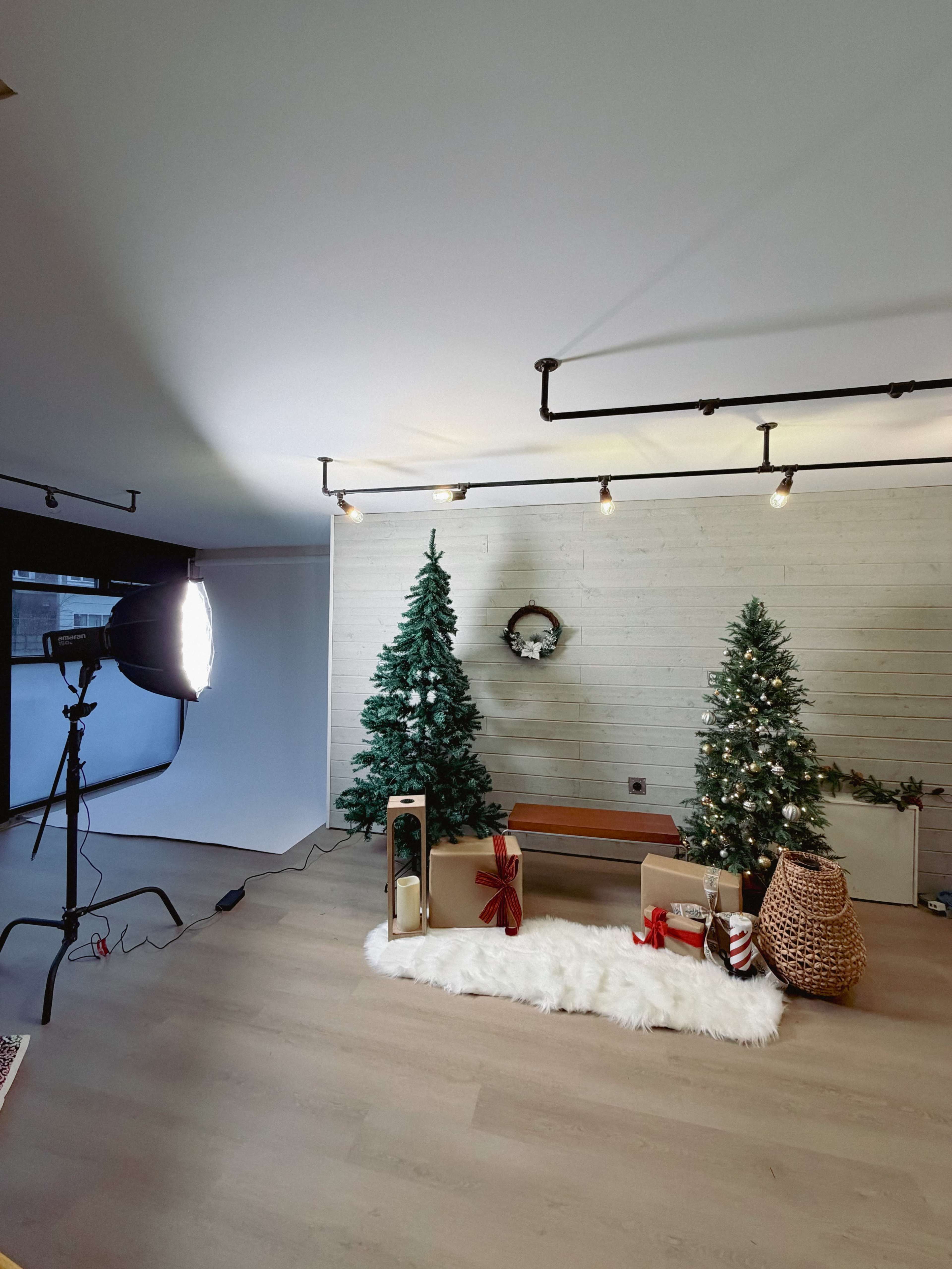 A cozy indoor setting features two Christmas trees, a wooden bench, a large basket, and wrapped gifts on a white fur rug, illuminated by soft lighting.