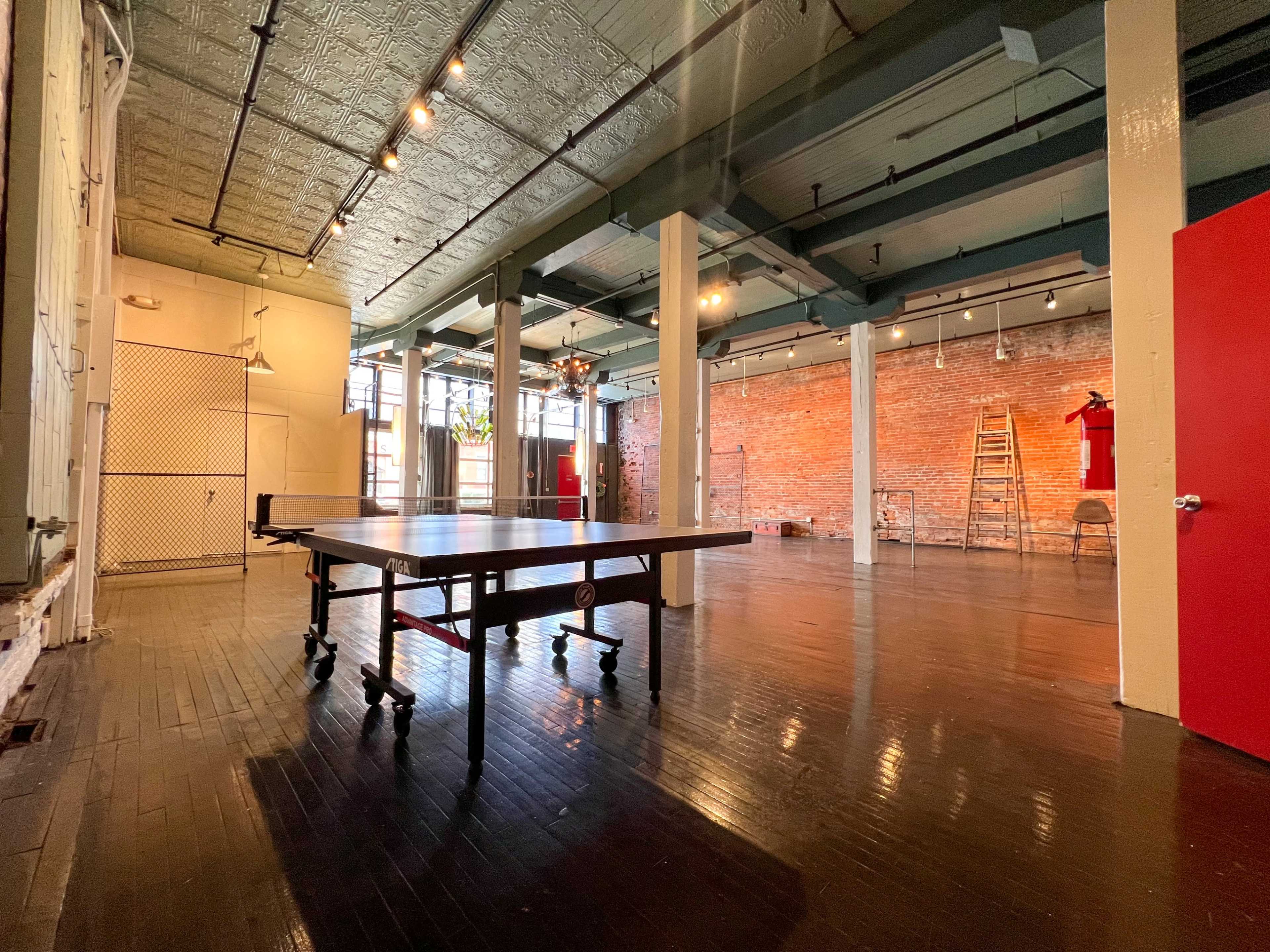 An industrial-style interior space featuring a large table in the center, brick walls, and various chairs and equipment in the background.