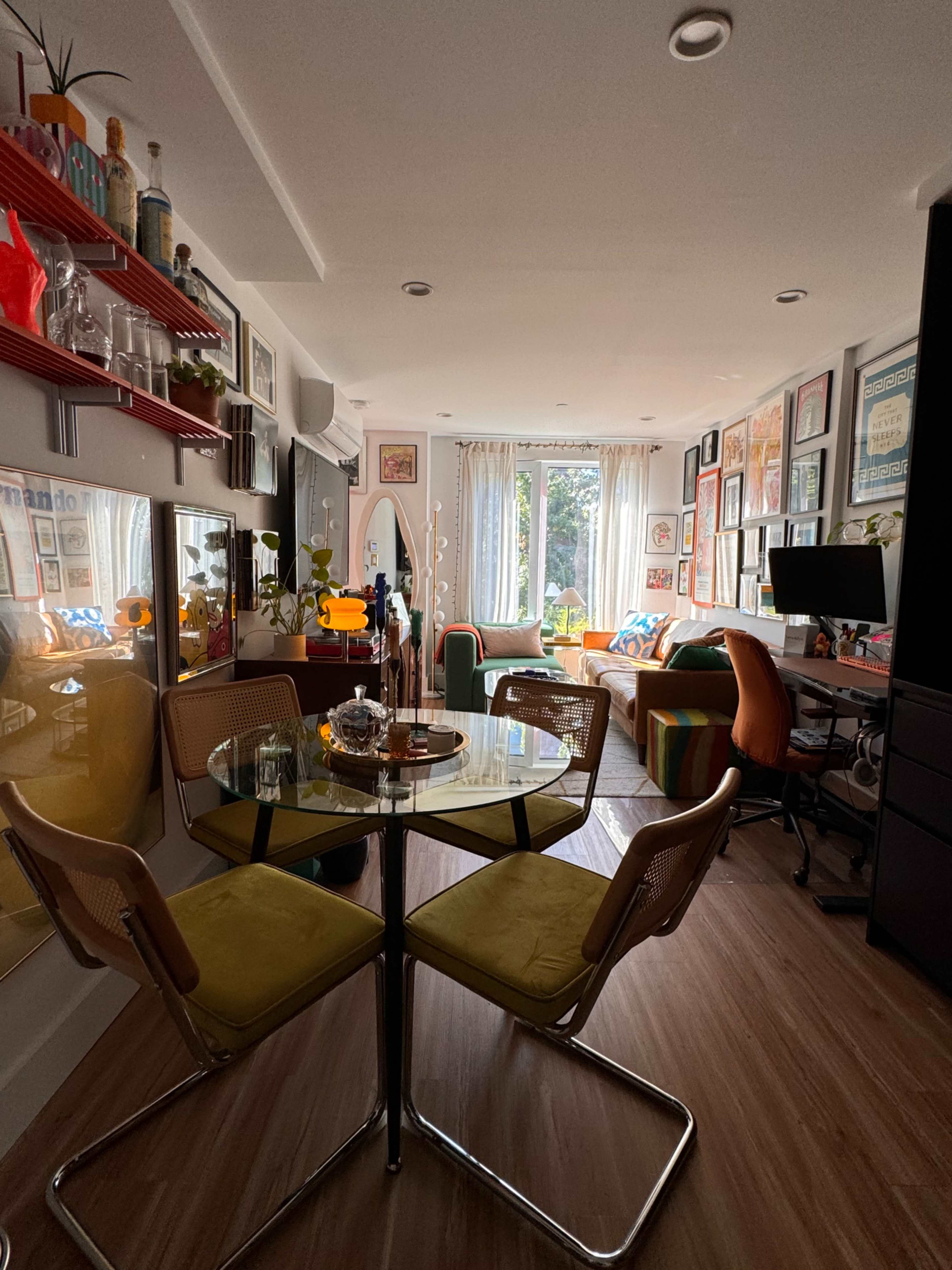 A round glass table with yellow chairs is positioned in the foreground of a brightly lit room filled with colorful decor and framed artwork.