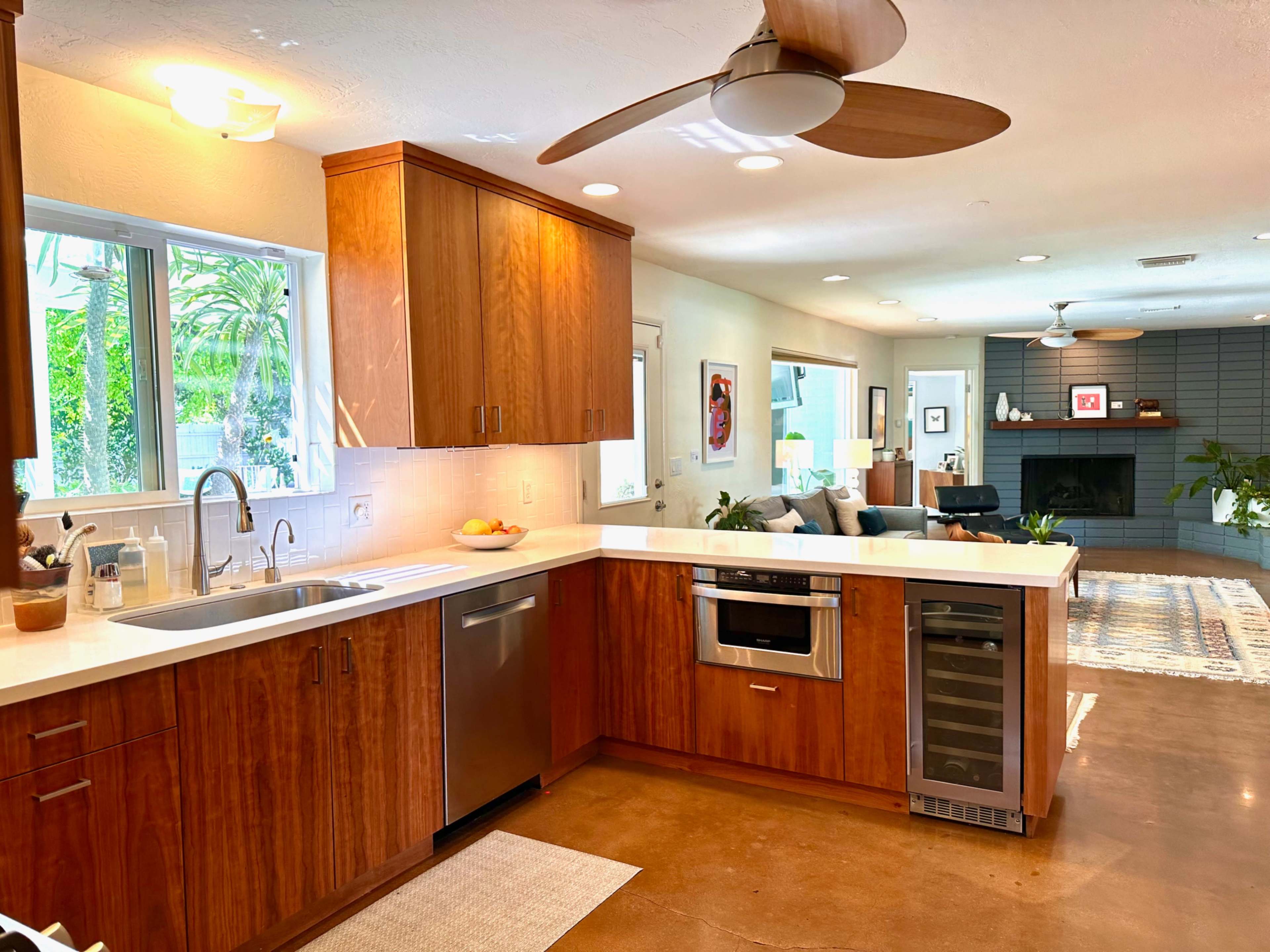 A modern kitchen with wooden cabinets, a stainless steel oven, and a white countertop, adjacent to a living area featuring a fireplace and seating.