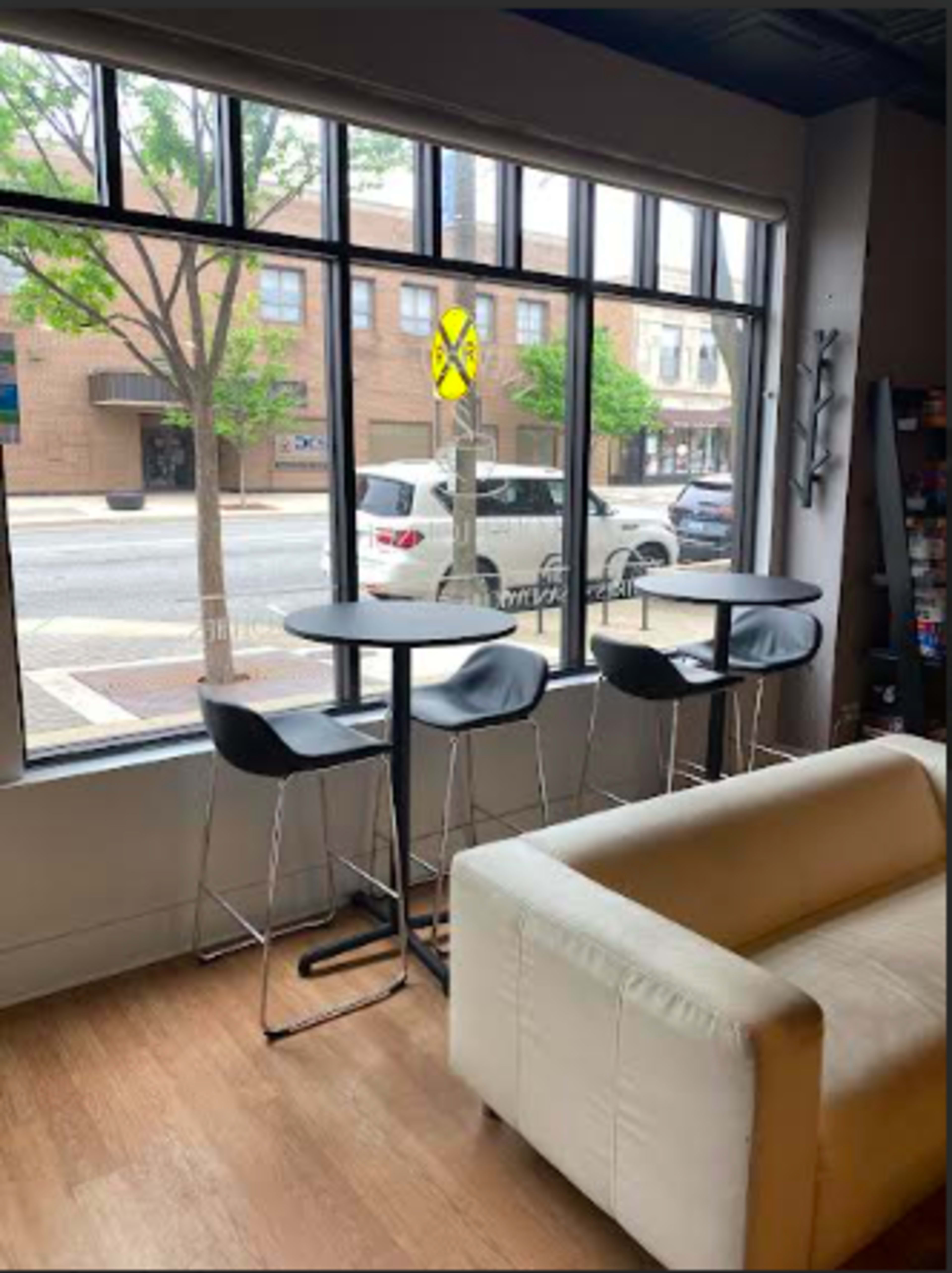 The image shows a small café interior with three black bar stools at a tall round table next to a large window overlooking a street with trees and parked cars.