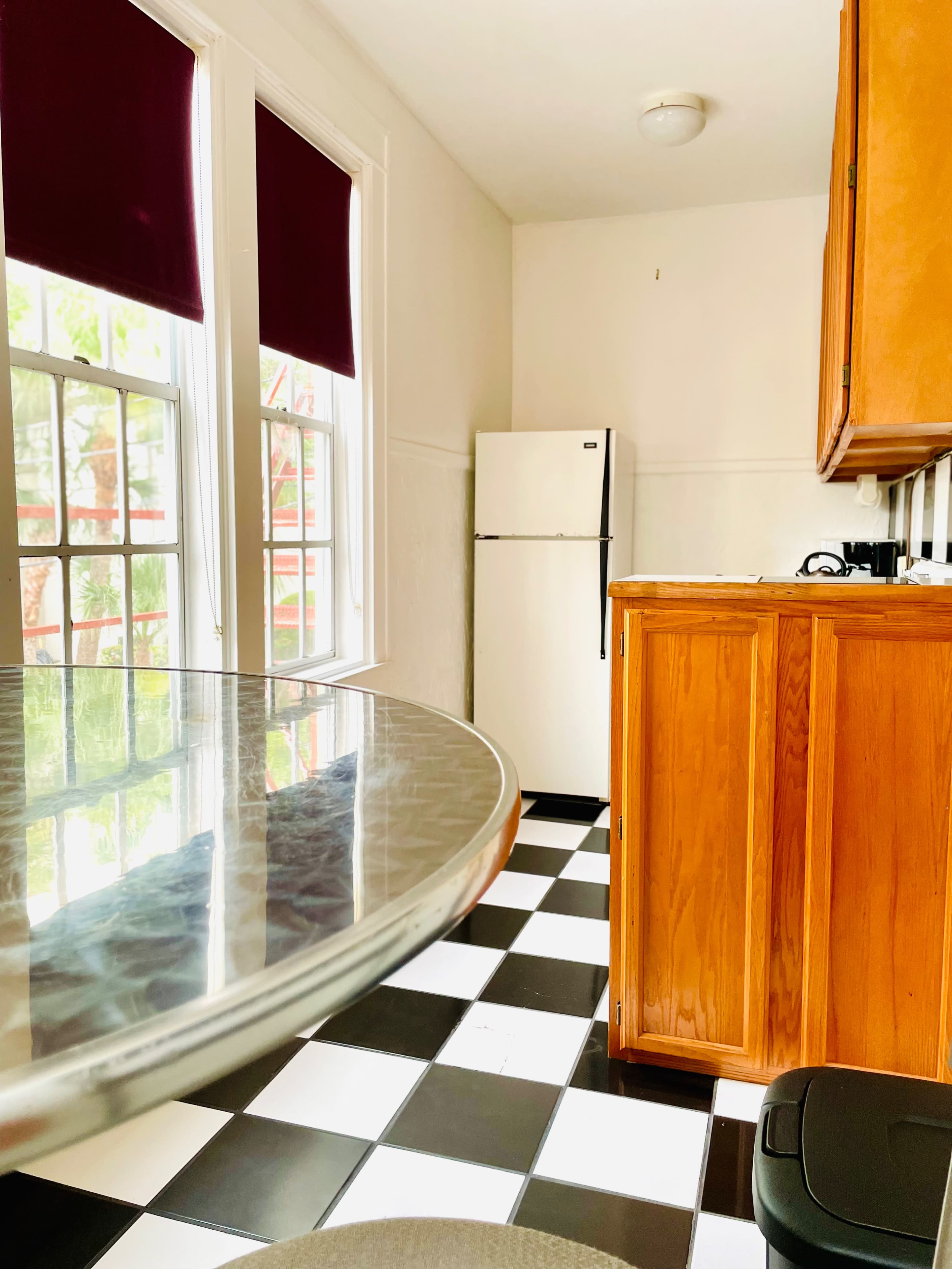 A kitchen with a round glass table in the foreground, a black and white checkered floor, and a white refrigerator against a wall with wooden cabinets.
