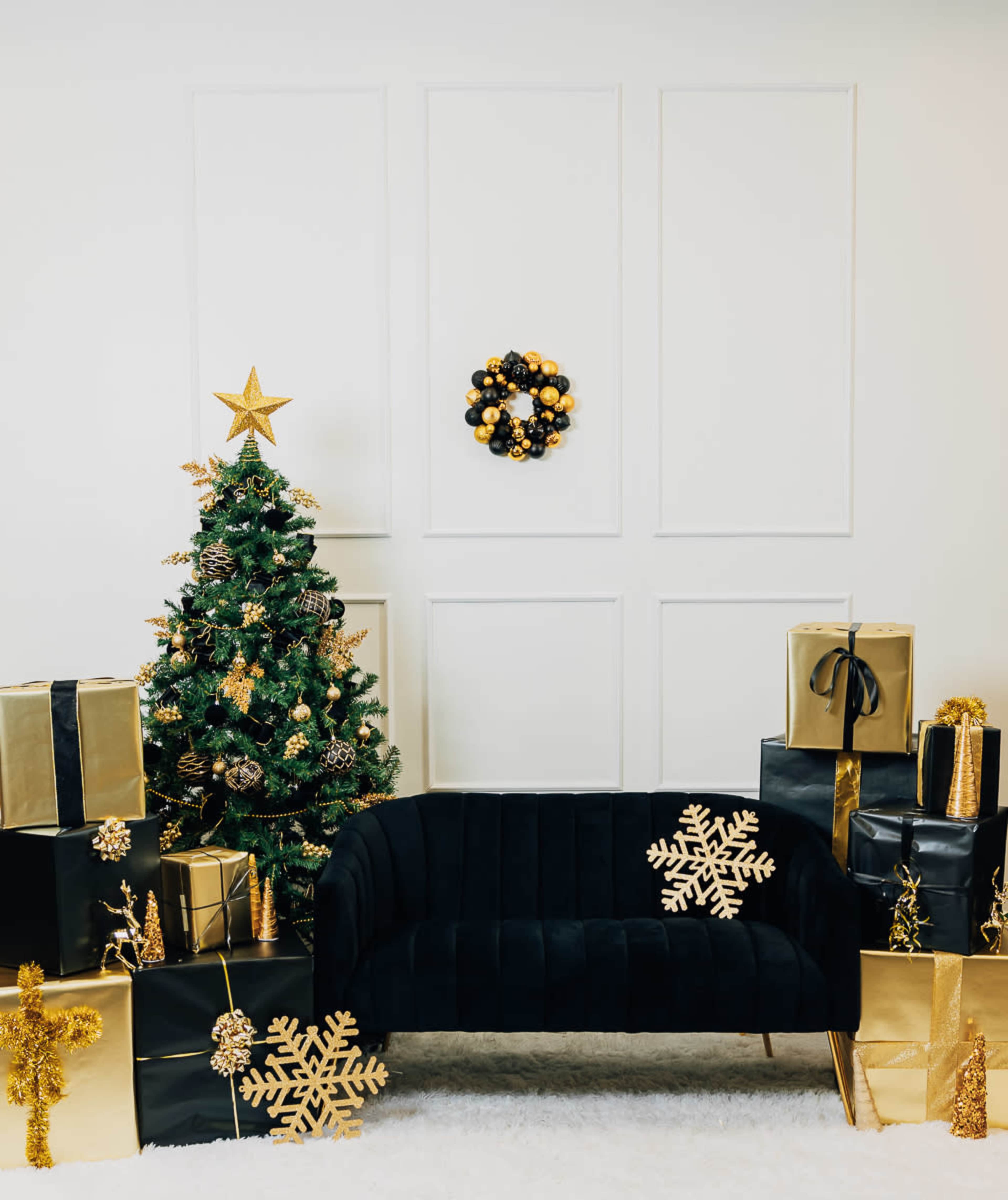 The scene features a decorated Christmas tree next to a black sofa, surrounded by wrapped gifts in gold and black, with a wreath on the wall.