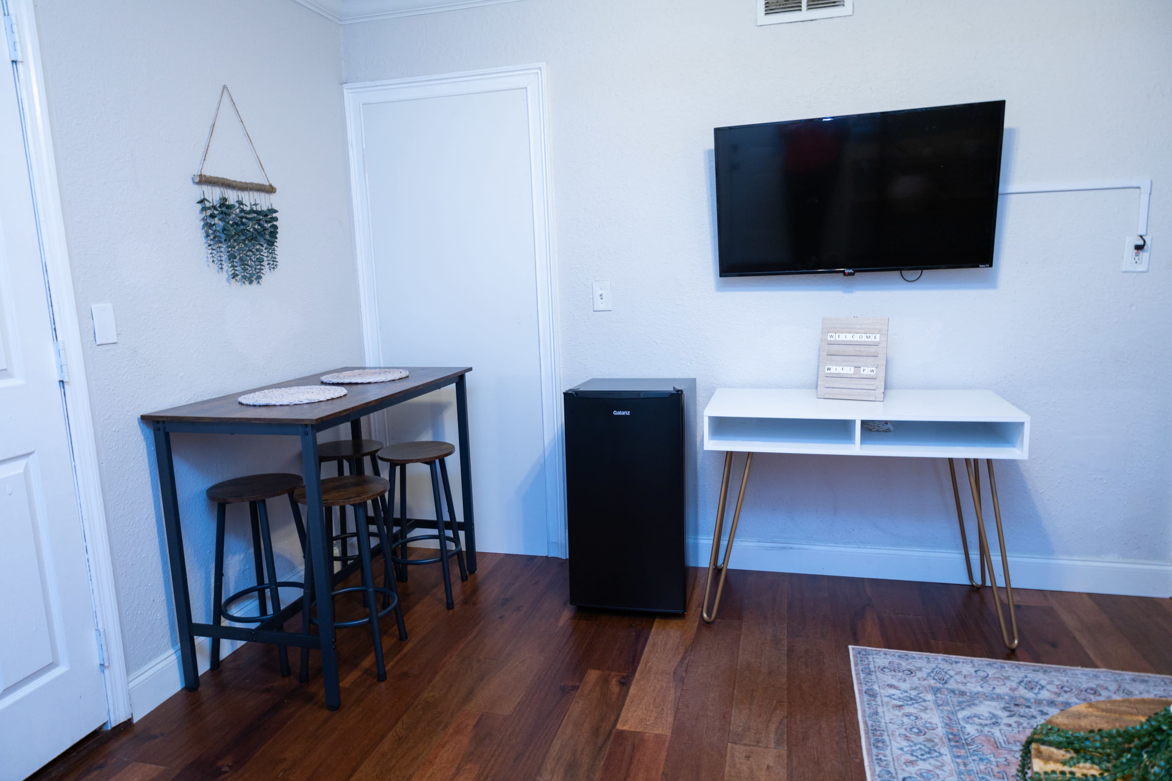 The image shows a small room with a table and three stools, a black mini fridge, and a wall-mounted television above a white console table.