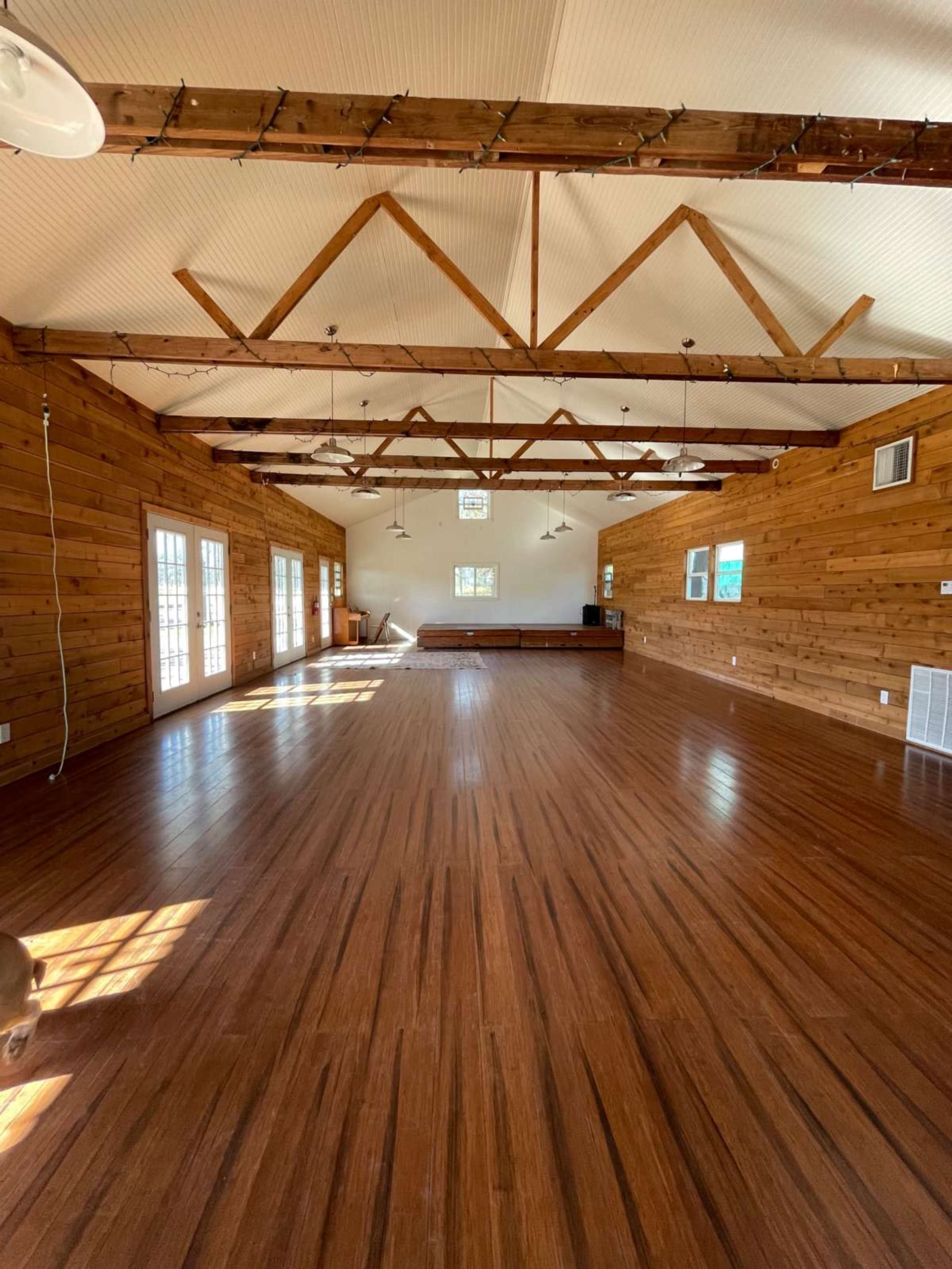 The image shows an empty wooden interior space with large windows and exposed beams, featuring a polished wooden floor.