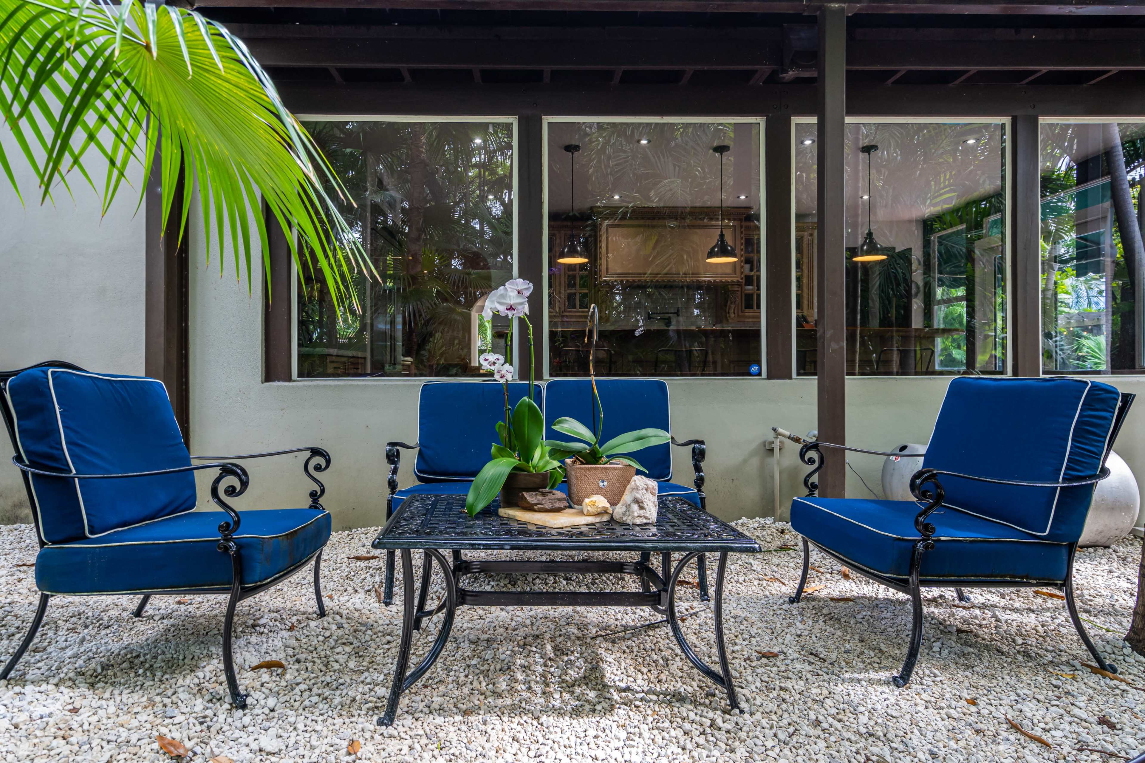 A patio area features four blue cushioned chairs surrounding a black metal coffee table, set on a bed of white gravel with greenery and large windows in the background.