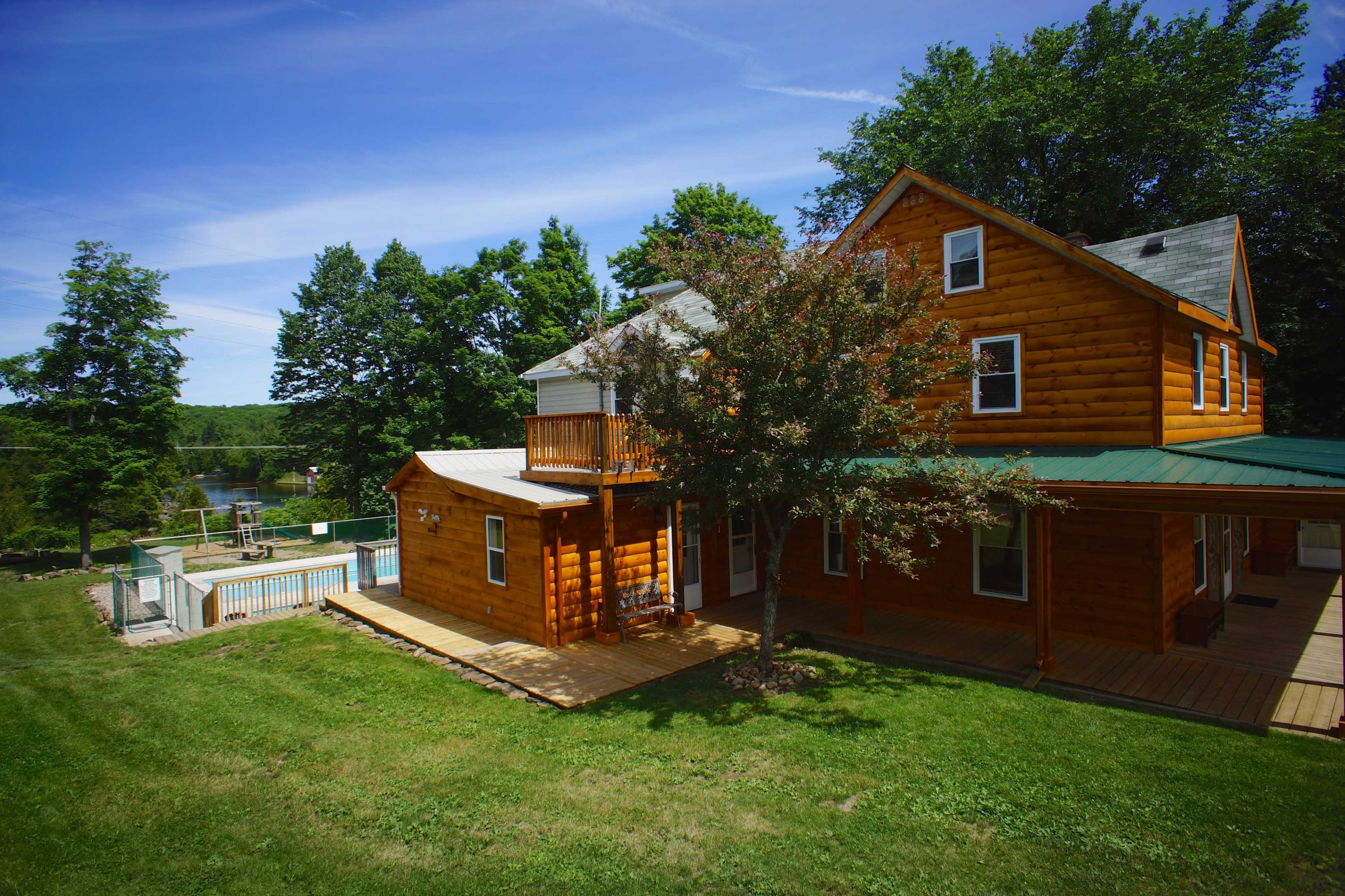 A large wooden house with a green roof and a deck overlooks a pool and a lake surrounded by trees.